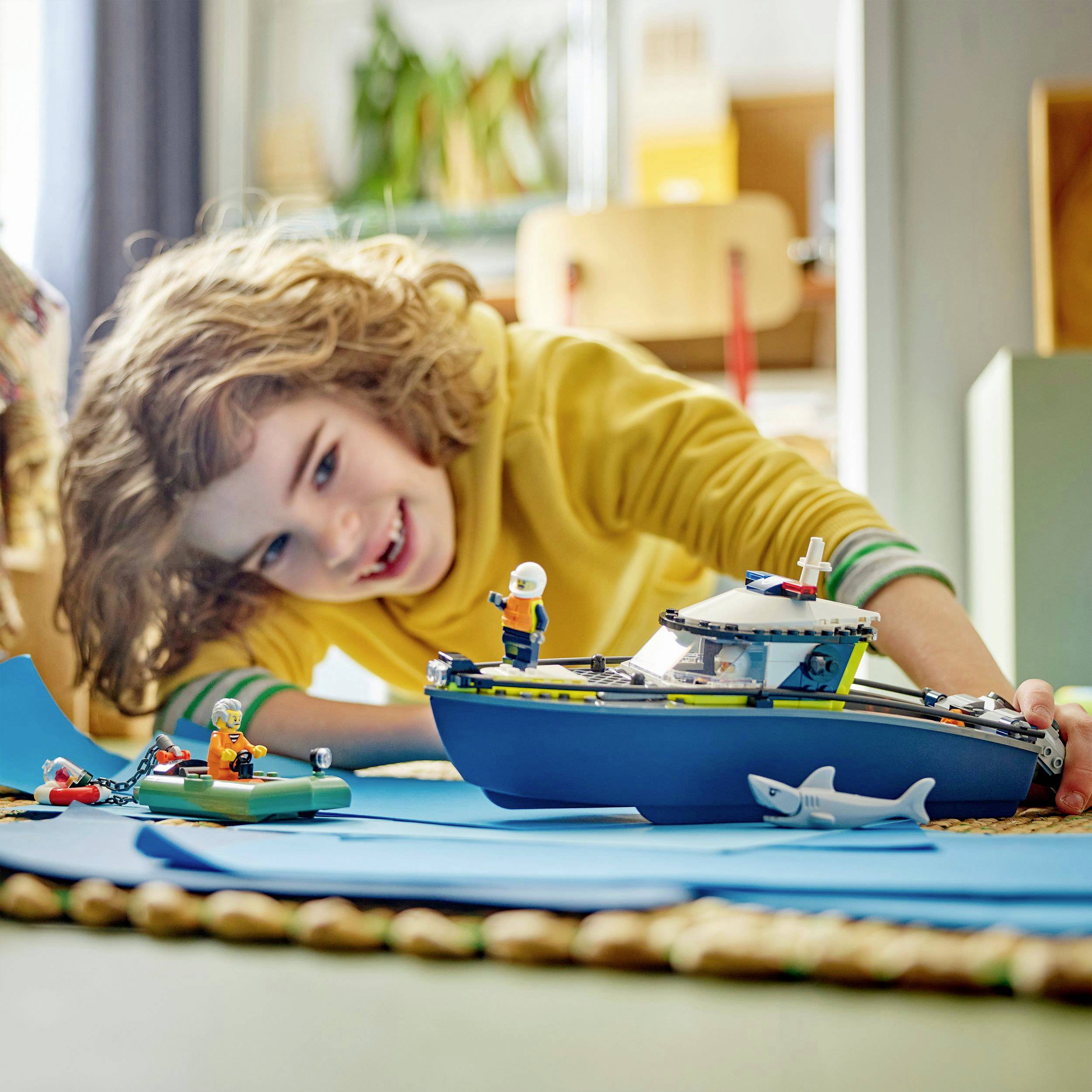 Un enfant joyeux joue avec une maquette de bateau et des figurines sur un tapis bleu. À l'arrière-plan, on peut voir des meubles et des plantes.