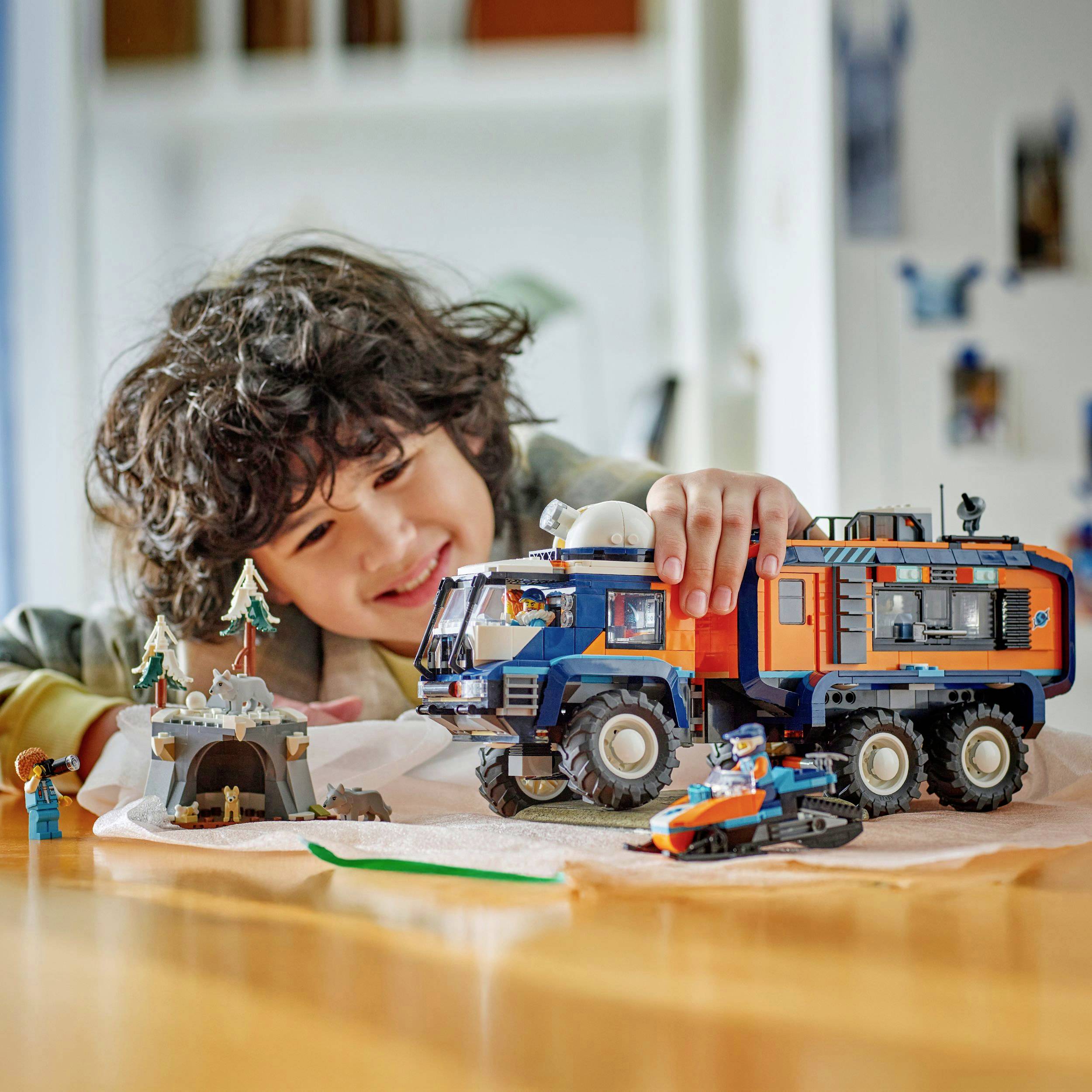 Un enfant joue en souriant avec des véhicules jouets et un château de sable sur une table dans un espace de jeu intérieur.