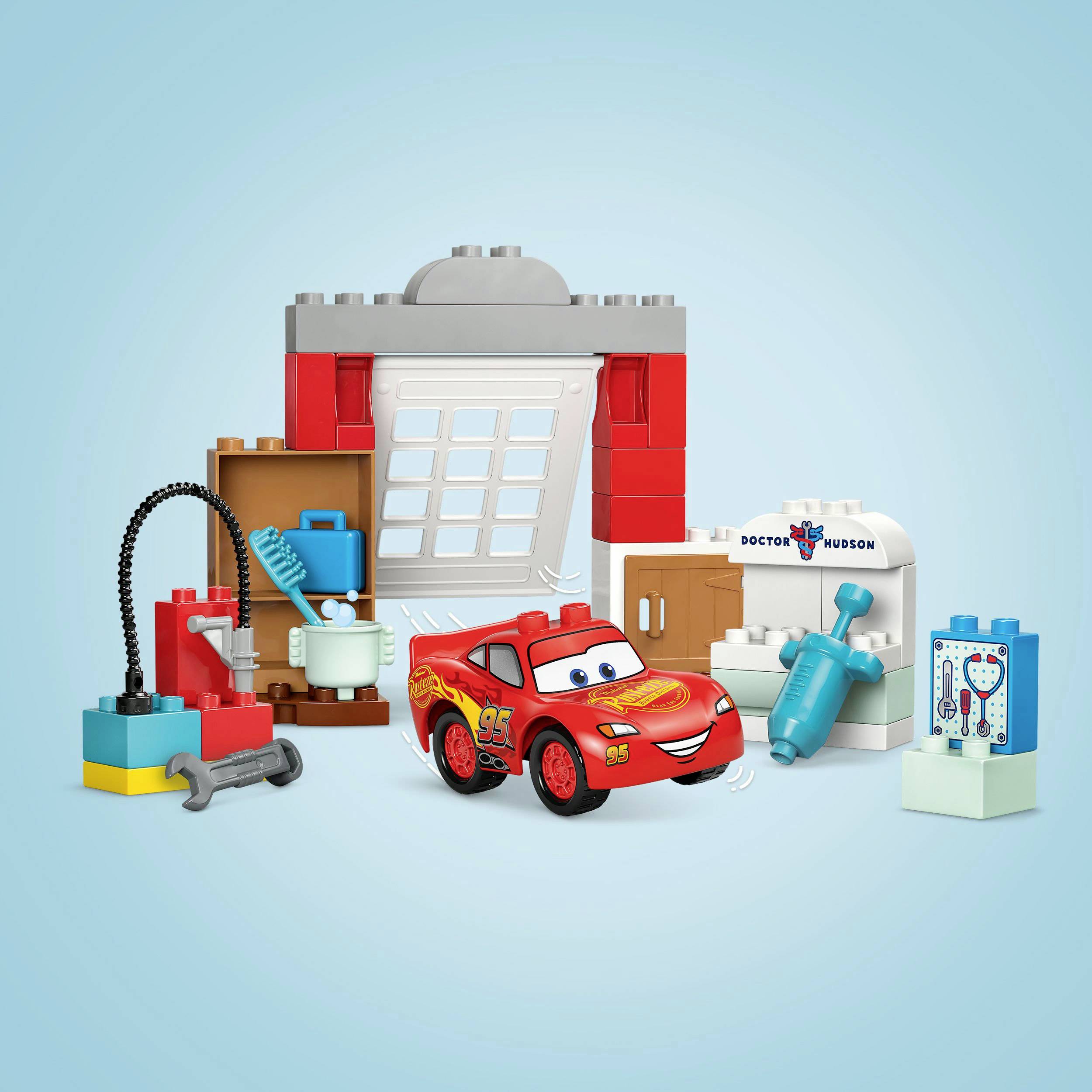 Une petite voiture rouge est garée devant un bâtiment fait de briques colorées. À côté se trouvent des outils et des accessoires.