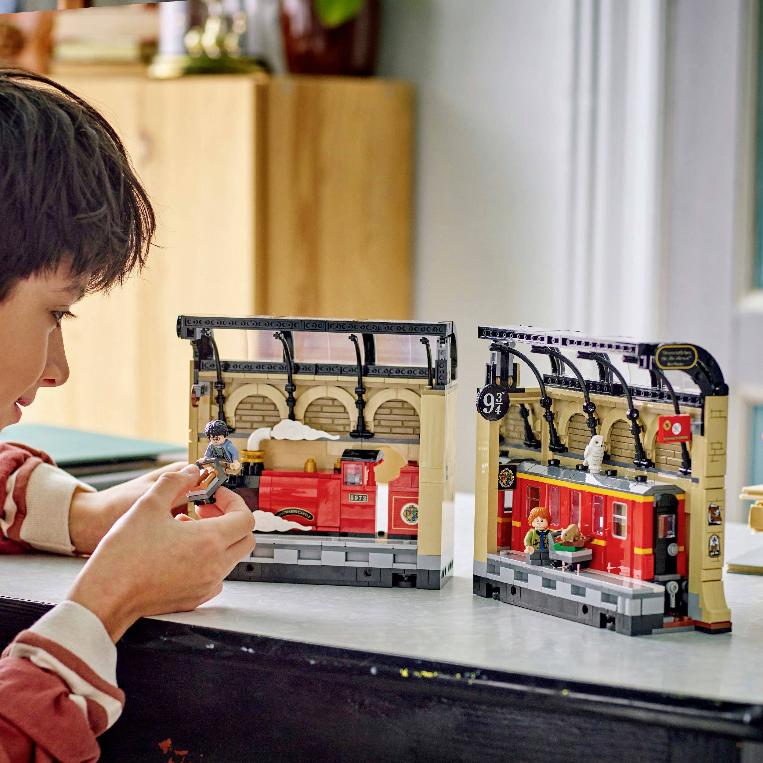 Un enfant joue avec une gare miniature qui évoque une scène magique. Le décor présente un modèle de train jouet rouge.