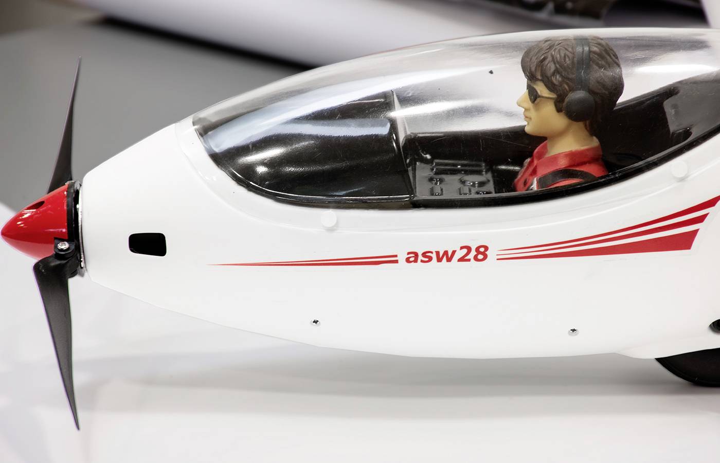 Un modèle réduit d'avion en gros plan, avec un cockpit détaillé et un modèle de pilote. Inscription 'asw28' et lignes rouges imprimées.