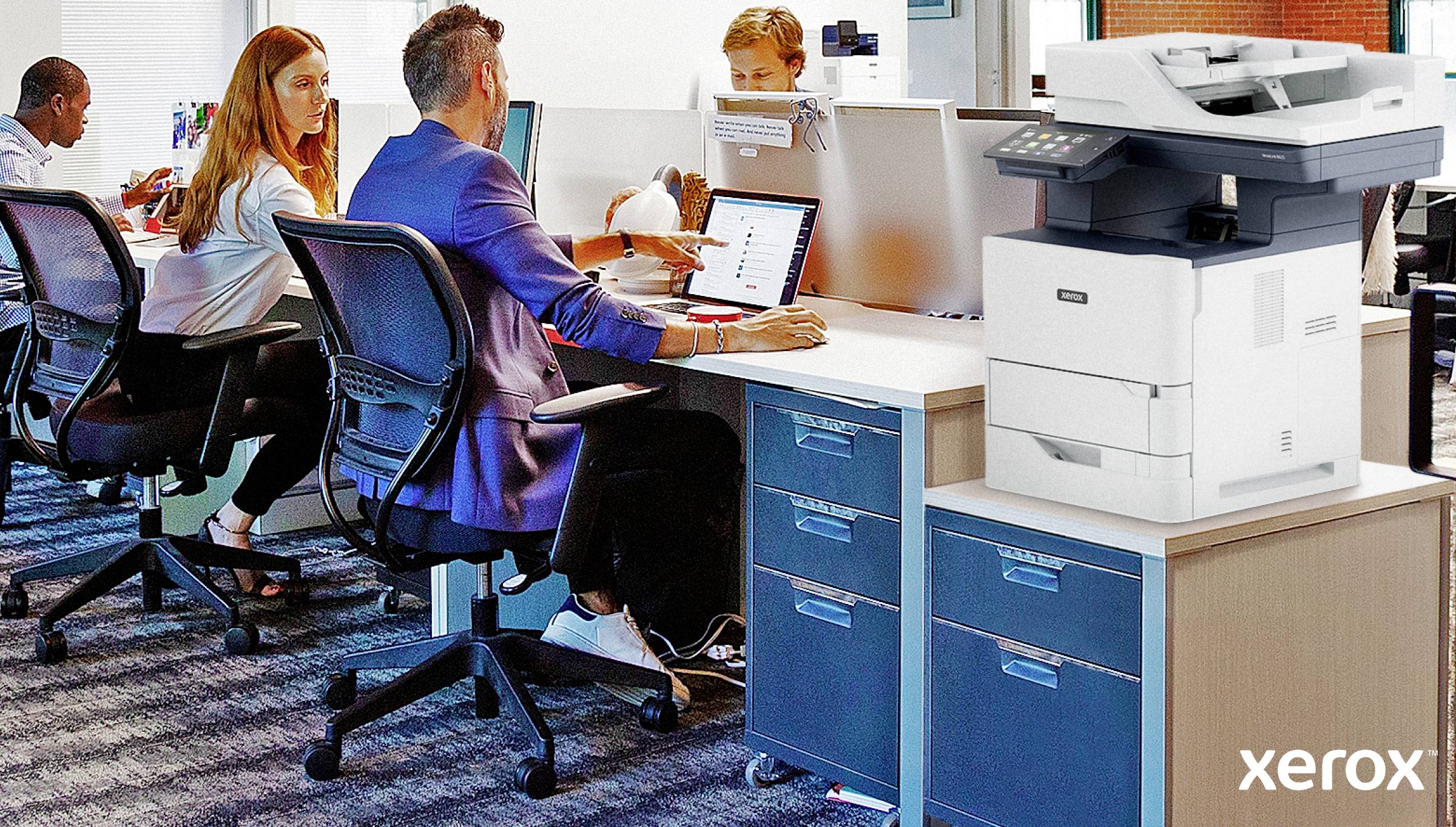 Des employés travaillent dans un bureau avec des ordinateurs. Une imprimante est posée sur une armoire au premier plan.