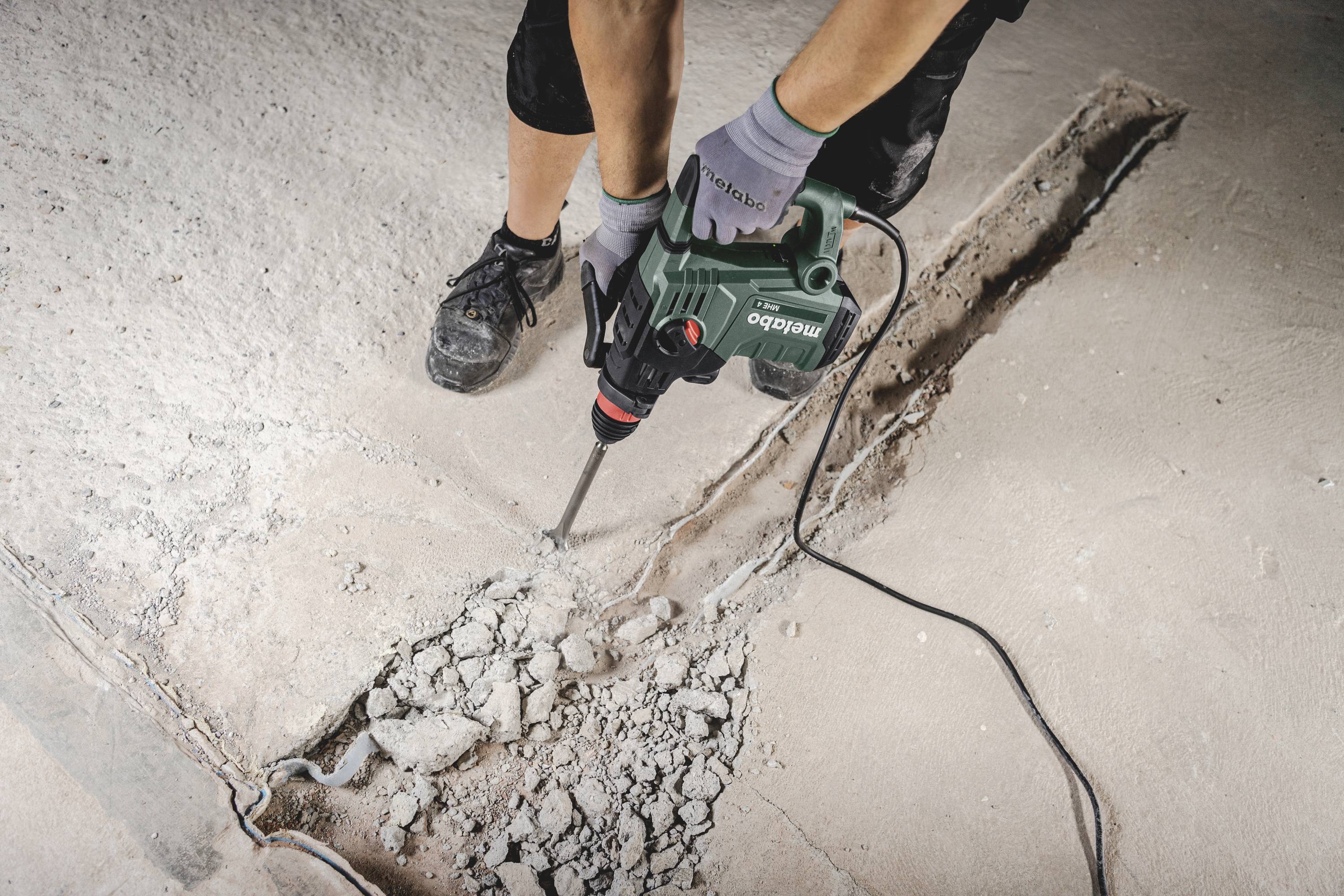 Personne utilisant un marteau-piqueur vert pour casser un sol en béton. Le sol est partiellement brisé, et des débris sont éparpillés tout autour.
