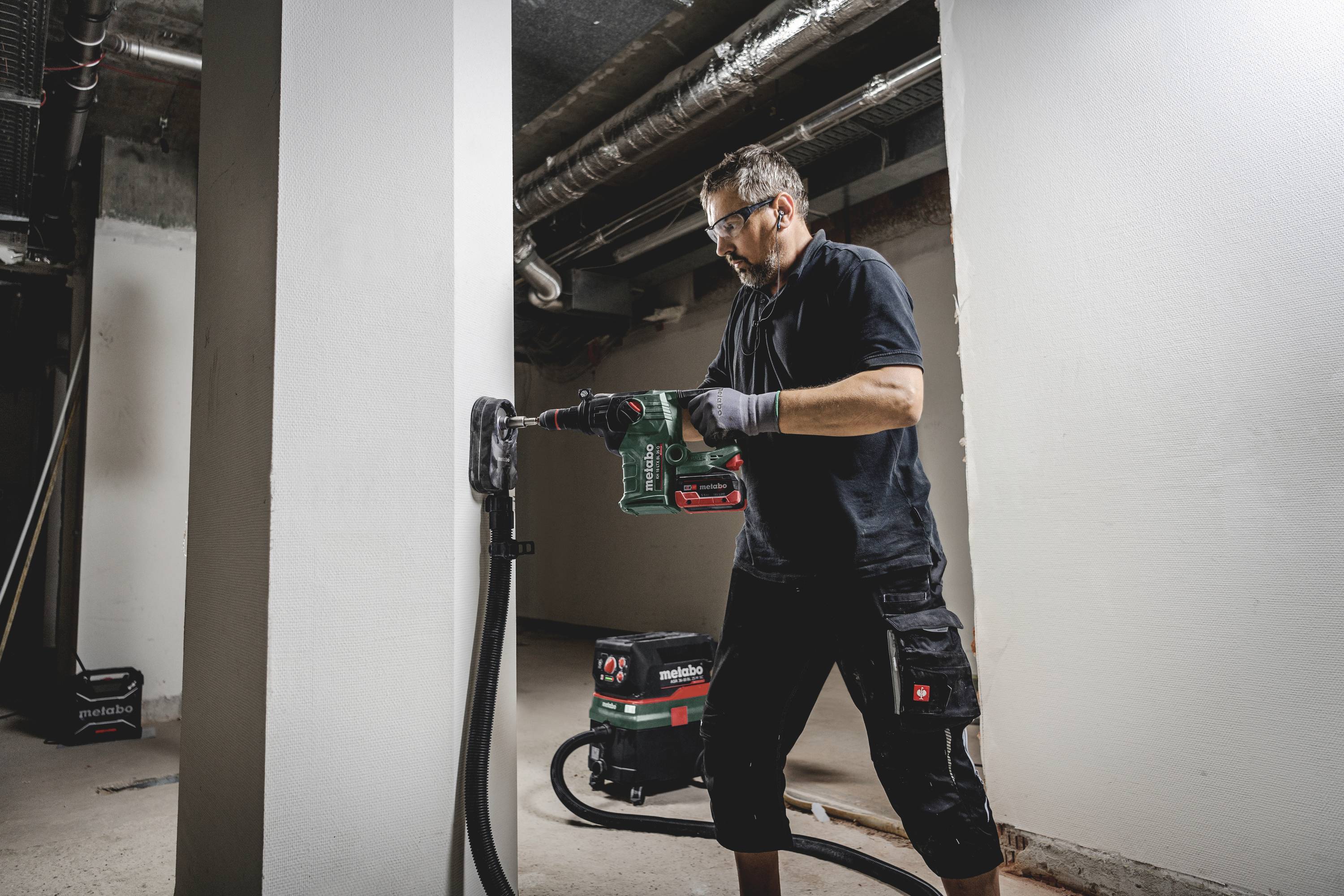 Un homme fore un mur en béton avec une perceuse à percussion dans un espace industriel. Une unité d'aspiration est placée à côté de lui.