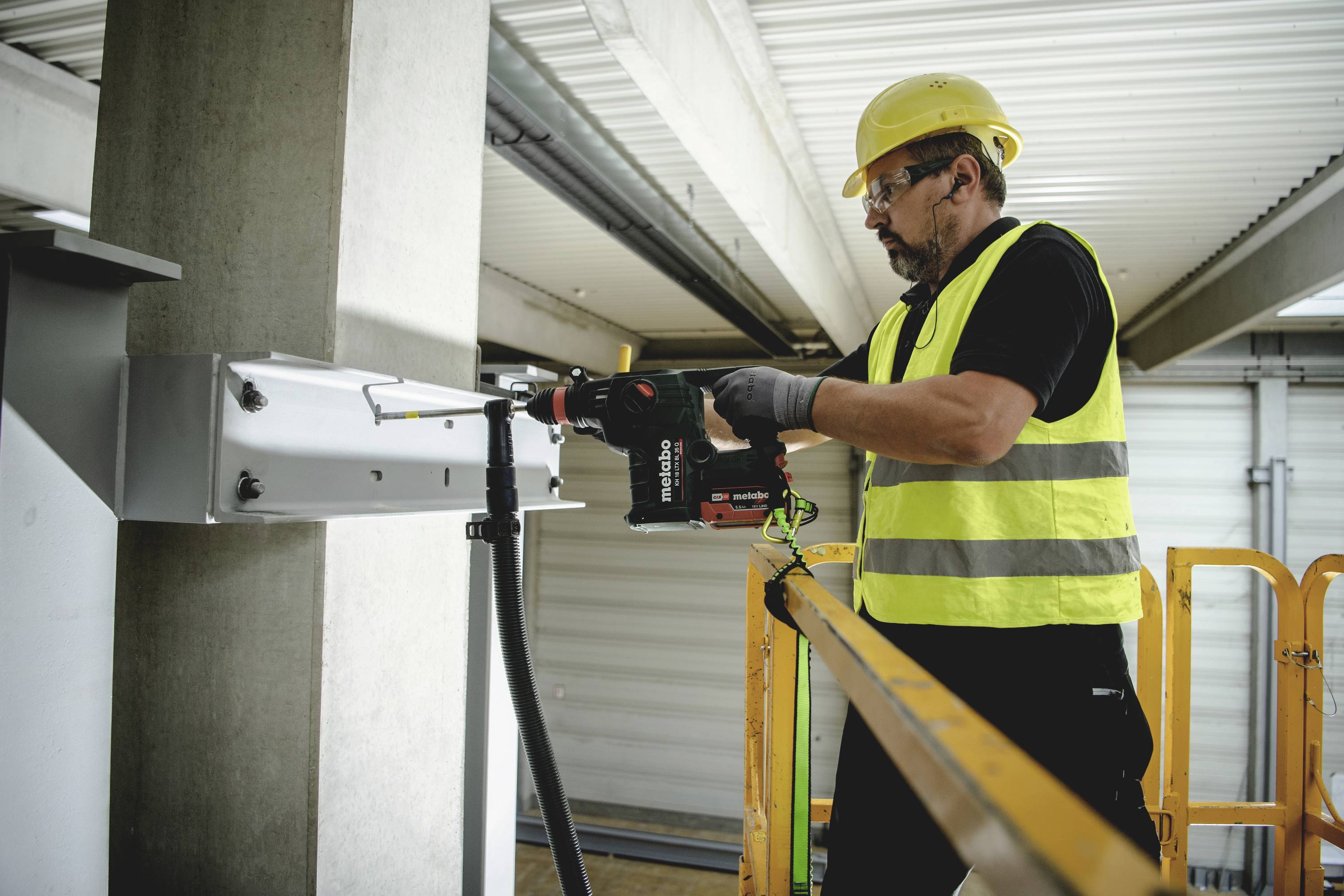 Un ouvrier portant un gilet jaune et un casque utilise une perceuse électrique pour travailler sur une structure métallique.
