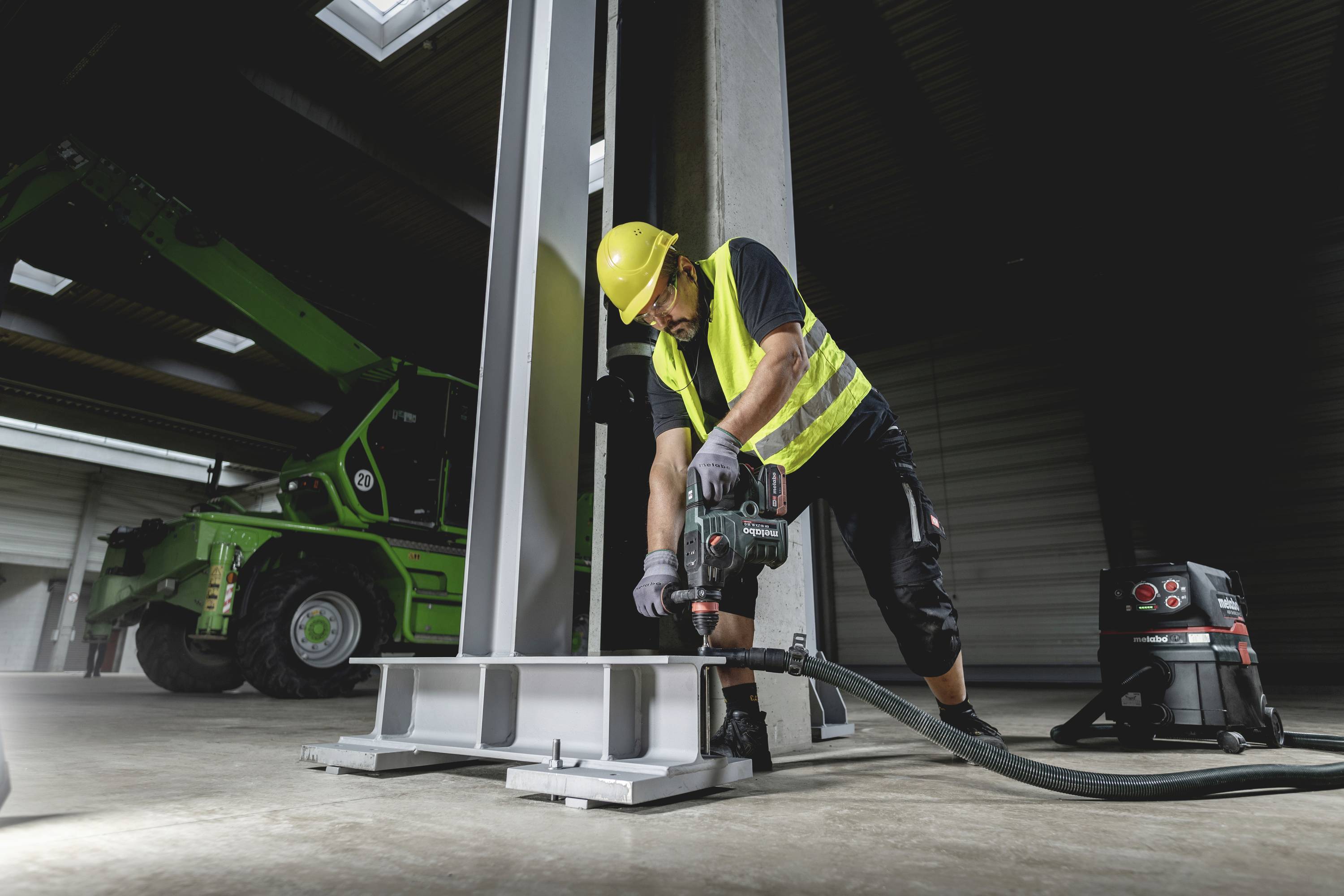 Un ouvrier portant un gilet jaune et un casque fore du métal dans un hall. Une grue verte est visible en arrière-plan.