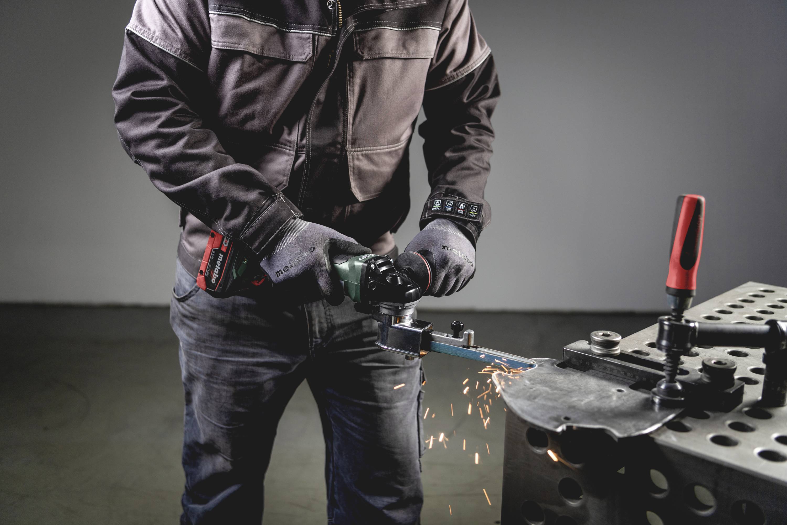 Une personne actionne une machine à meuler et génère des étincelles sur une table métallique. Travaille de manière concentrée dans un environnement d'atelier.