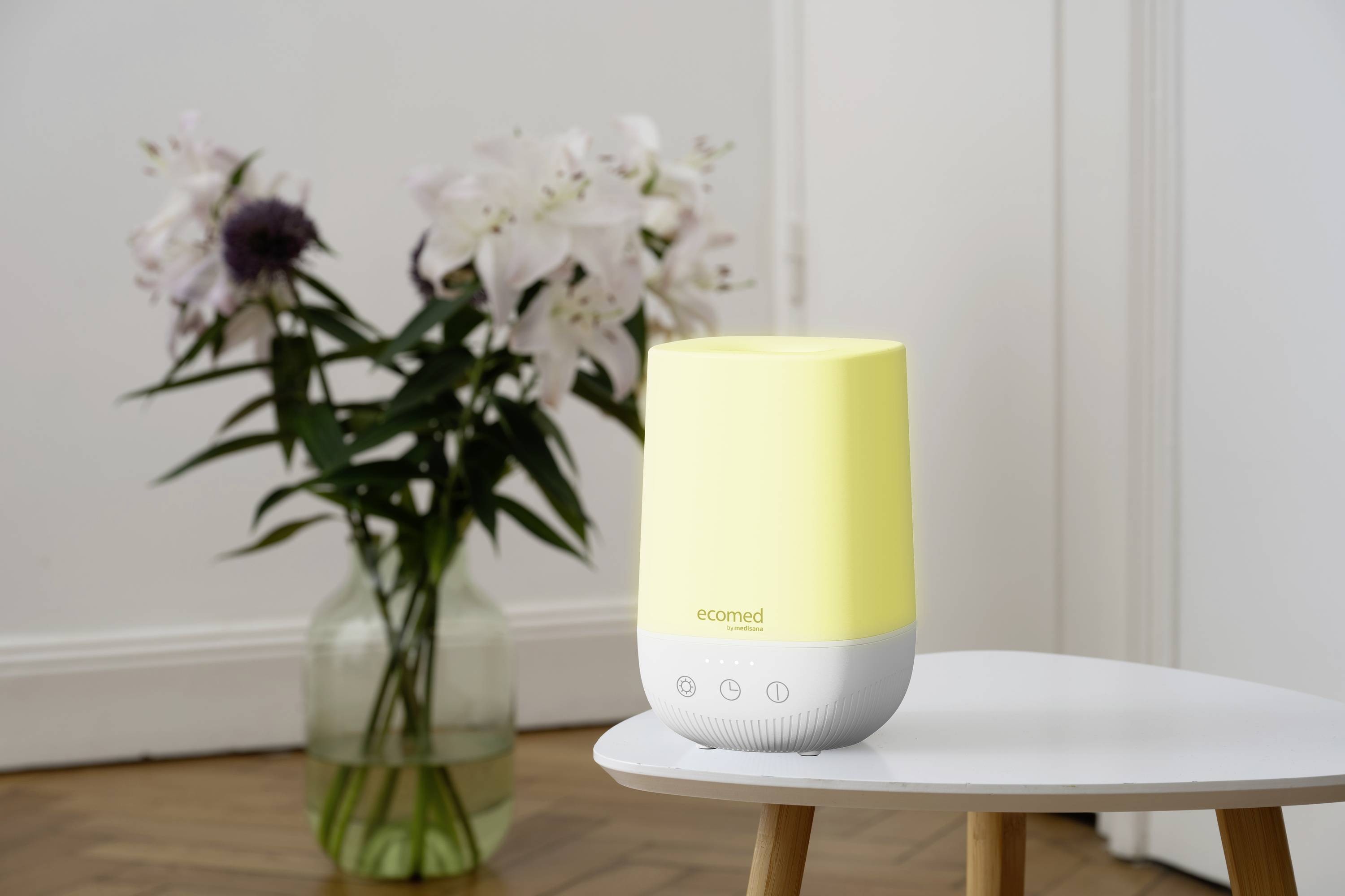 Un humidificateur jaune sur une table devant un bouquet de fleurs dans un vase, capturé dans une pièce lumineuse avec un sol en bois.
