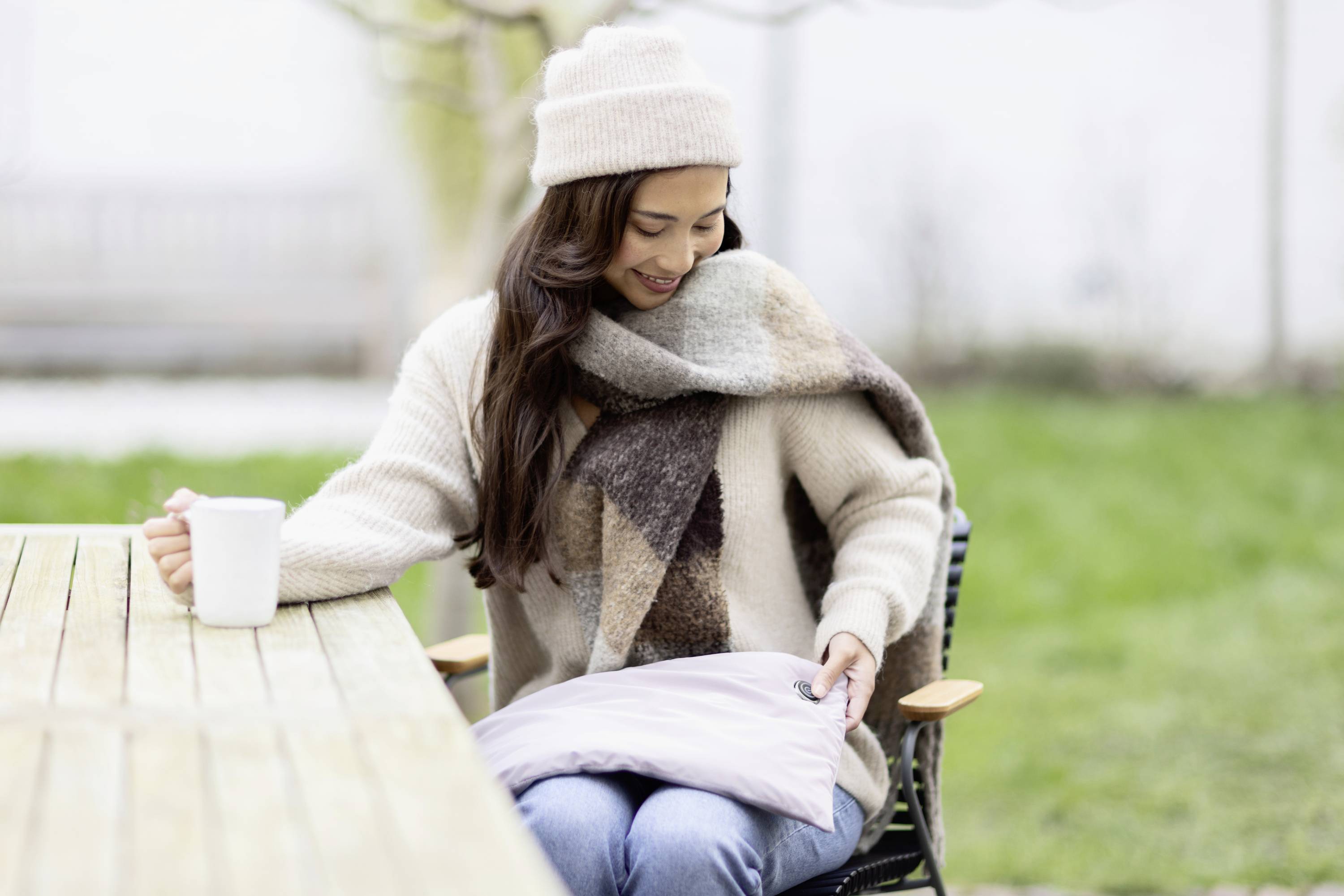 Une femme est assise dehors à une table en bois, tenant une tasse dans sa main droite et regardant une veste chaude sur ses genoux.