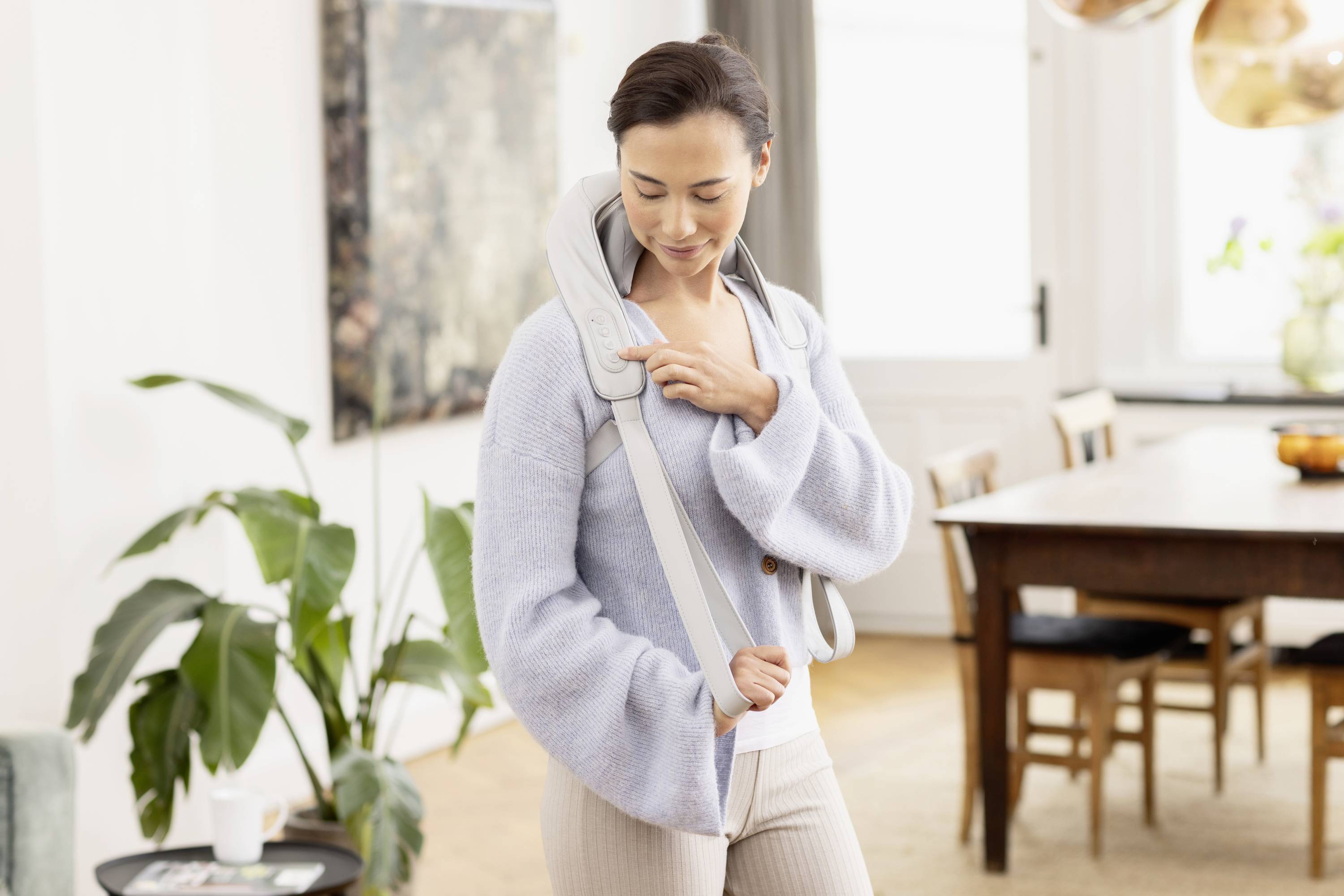 Une femme se tient dans une pièce lumineuse et utilise un appareil de massage cervical. Des plantes et une table sont visibles en arrière-plan.