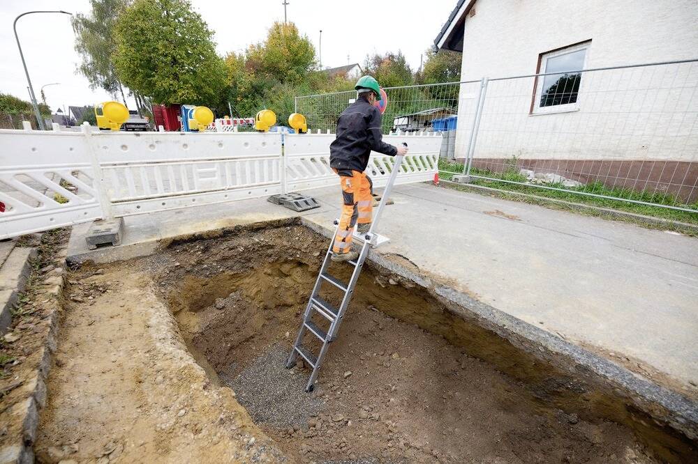 Une personne monte dans un grand trou creusé dans le sol sur un chantier à l'aide d'une échelle. Des clôtures de sécurité et des barrières entourent l'endroit.