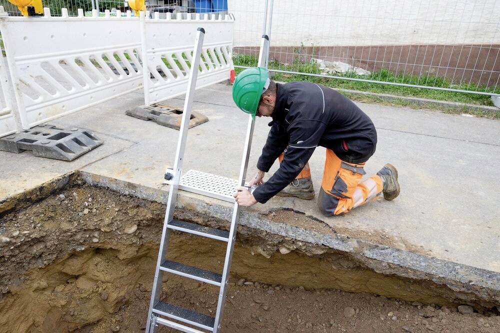 Un ouvrier portant un casque est agenouillé sur un chantier et fixe une échelle au bord d'une tranchée pour assurer un accès sécurisé à l'intérieur.