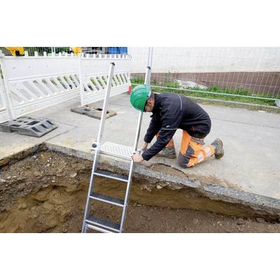 Un ouvrier portant un casque est agenouillé sur un chantier et fixe une échelle au bord d'une tranchée pour assurer un accès sécurisé à l'intérieur.