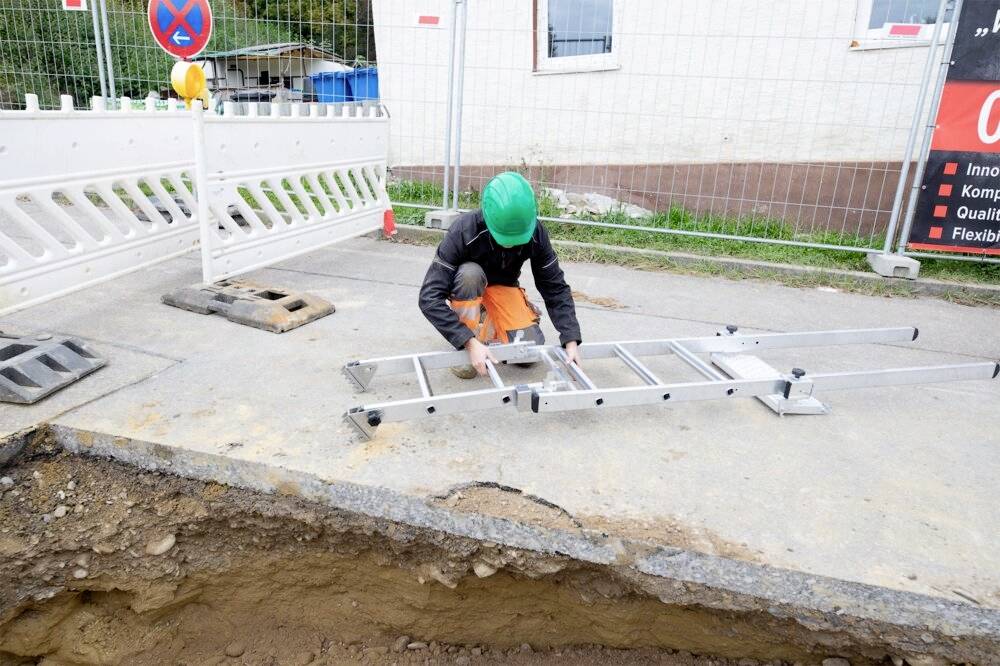 Une personne portant un casque de protection vert installe une échelle sur un chantier à côté d'une tranchée. Une clôture de chantier est visible en arrière-plan.