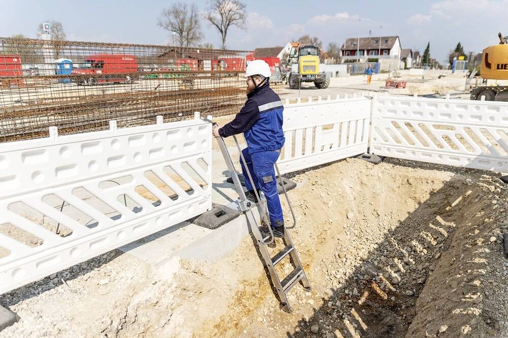 Un ouvrier portant un casque et une combinaison bleue descend une échelle dans une tranchée de chantier. Des équipements de construction et des barrières sont visibles en arrière-plan.