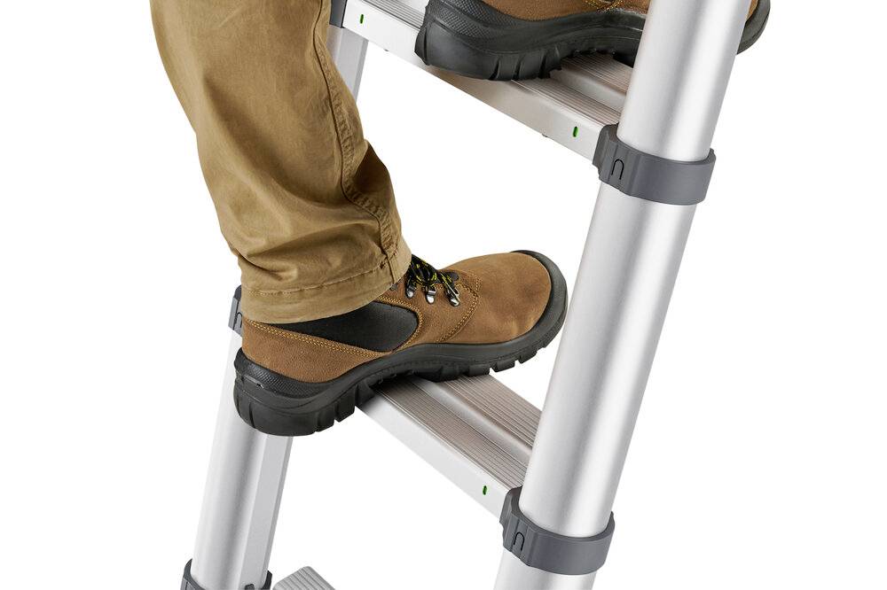 Une personne monte une échelle en aluminium, portant des chaussures brunes et un pantalon beige. Le focus est mis sur les pieds.