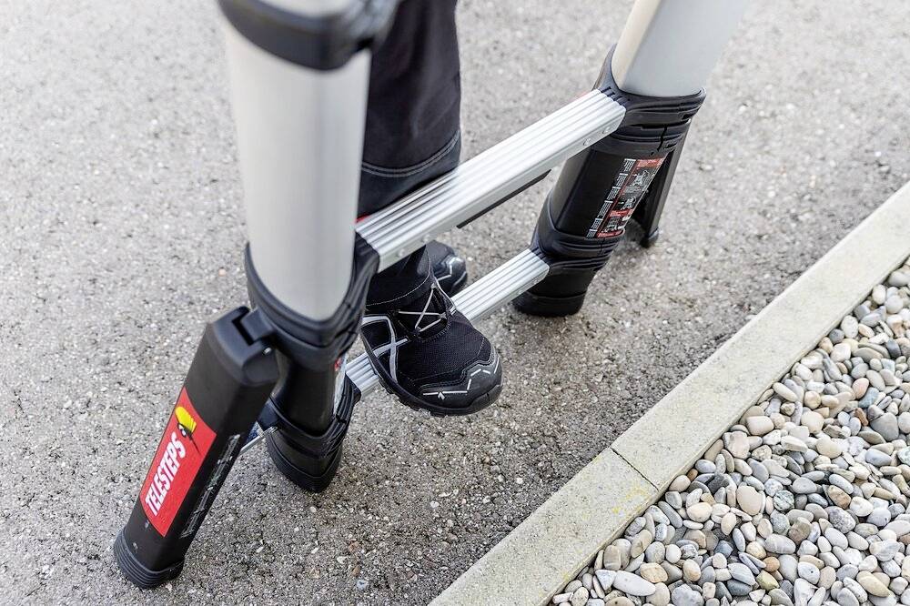 Une personne portant un pantalon sombre et des chaussures noires se tient sur une échelle télescopique. L'échelle est installée sur un chemin de gravier.