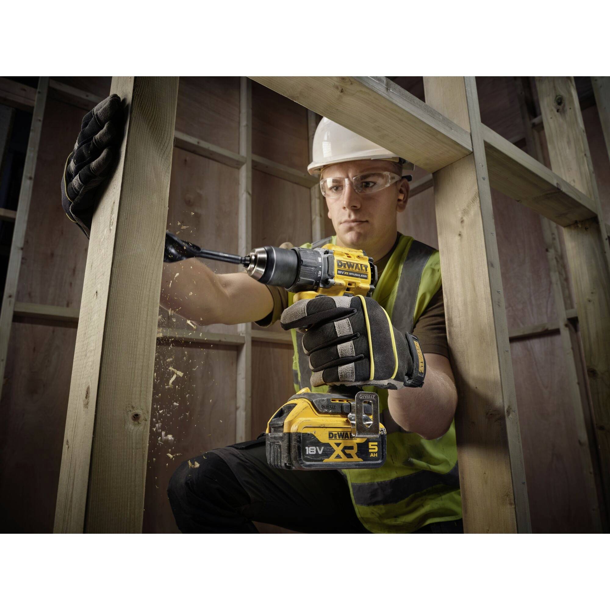 Un ouvrier porte des vêtements de protection et fore un cadre en bois à l'aide d'une perceuse sans fil dans un environnement de chantier.