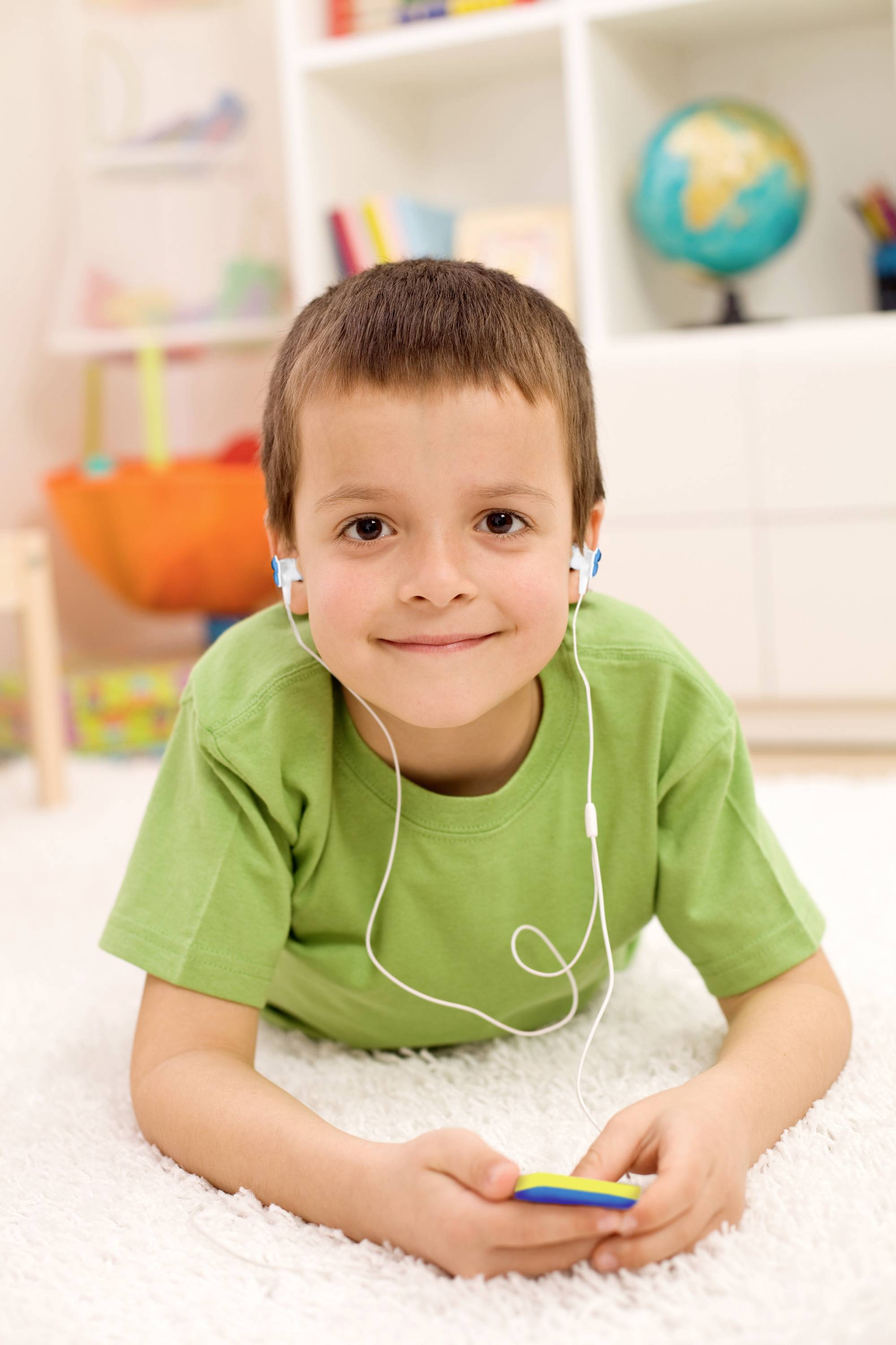 Un enfant est allongé sur un tapis, écoute de la musique avec des écouteurs et tient un appareil. En arrière-plan, on peut voir une étagère remplie de jouets et de livres.