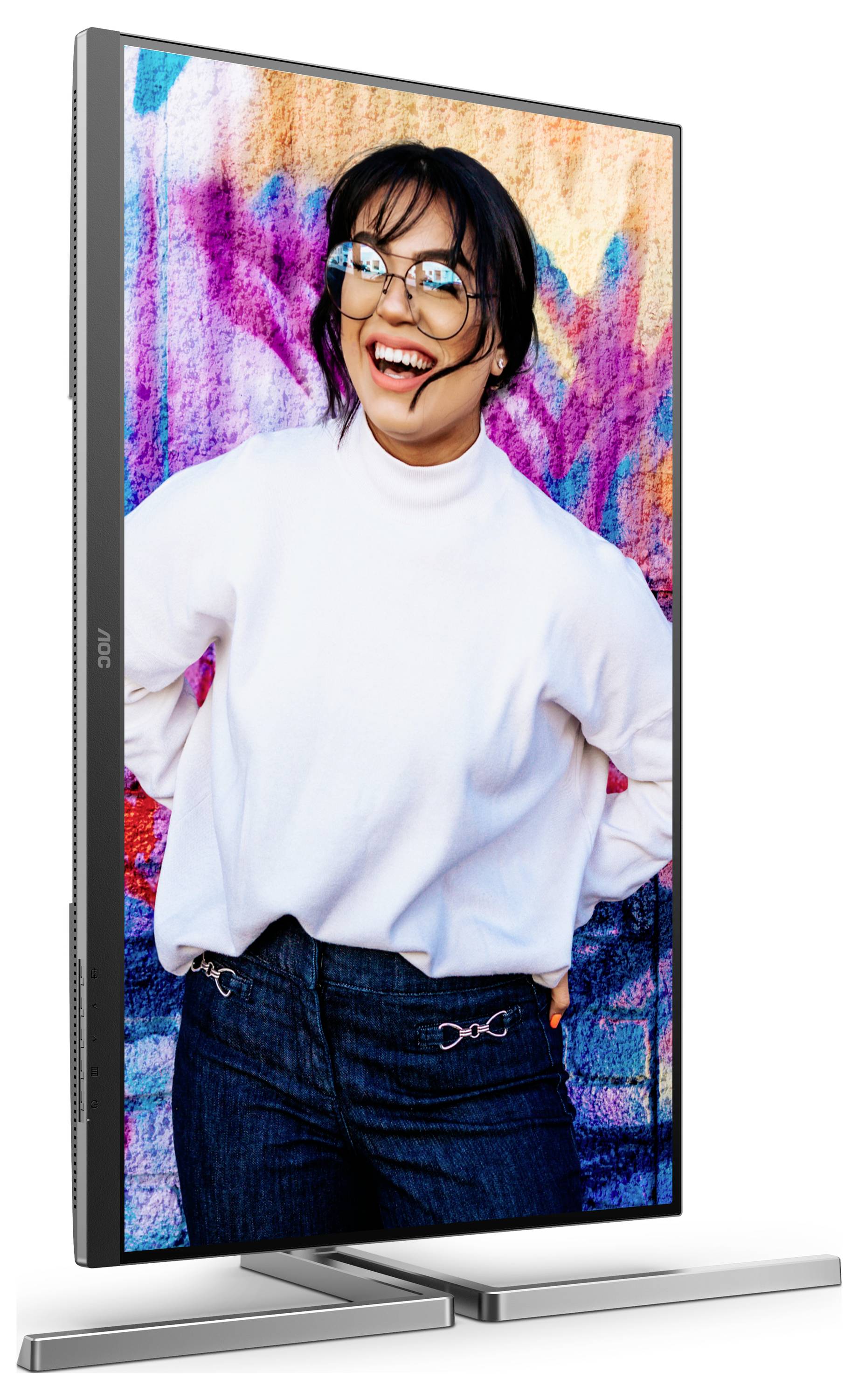 Une personne souriante devant un mur coloré et tagué. La personne porte des lunettes, un pull blanc et un jean foncé.