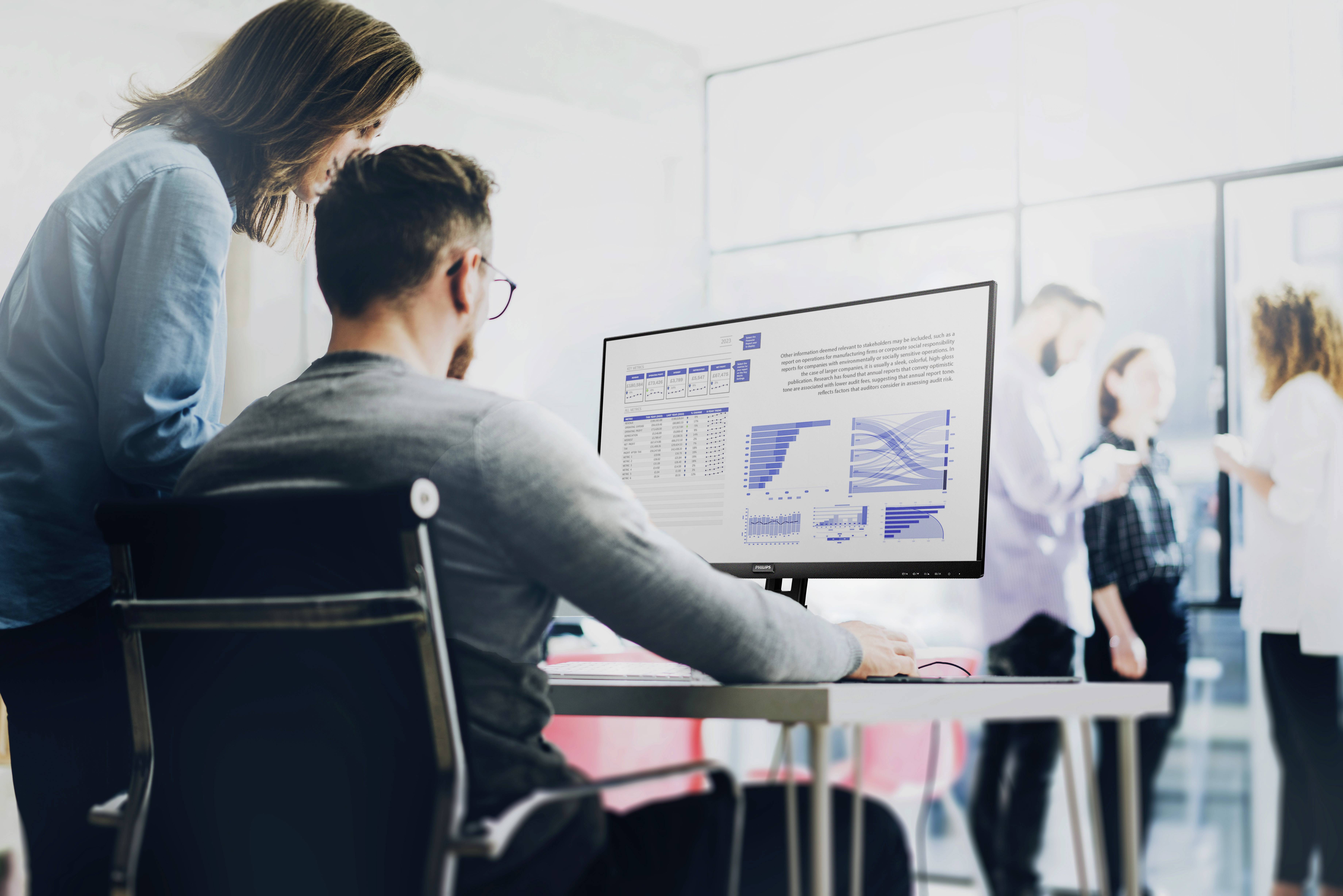 Une personne est assise à un bureau et examine des graphiques et des tableaux sur un écran. Une autre personne se tient à côté et regarde.