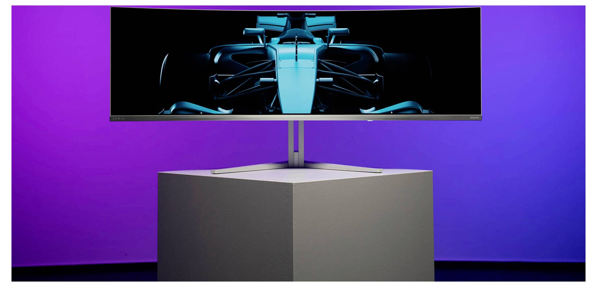 Un moniteur ultra-large affiche l'image d'une voiture de course futuriste. Le moniteur est posé sur un piédestal blanc devant un fond lilas.