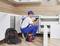 Un homme à tout faire portant une salopette bleue et une casquette assemble des armoires de cuisine à l'aide d'une perceuse électrique. Un sac à outils contenant des outils rouges et jaunes est posé à proximité.
