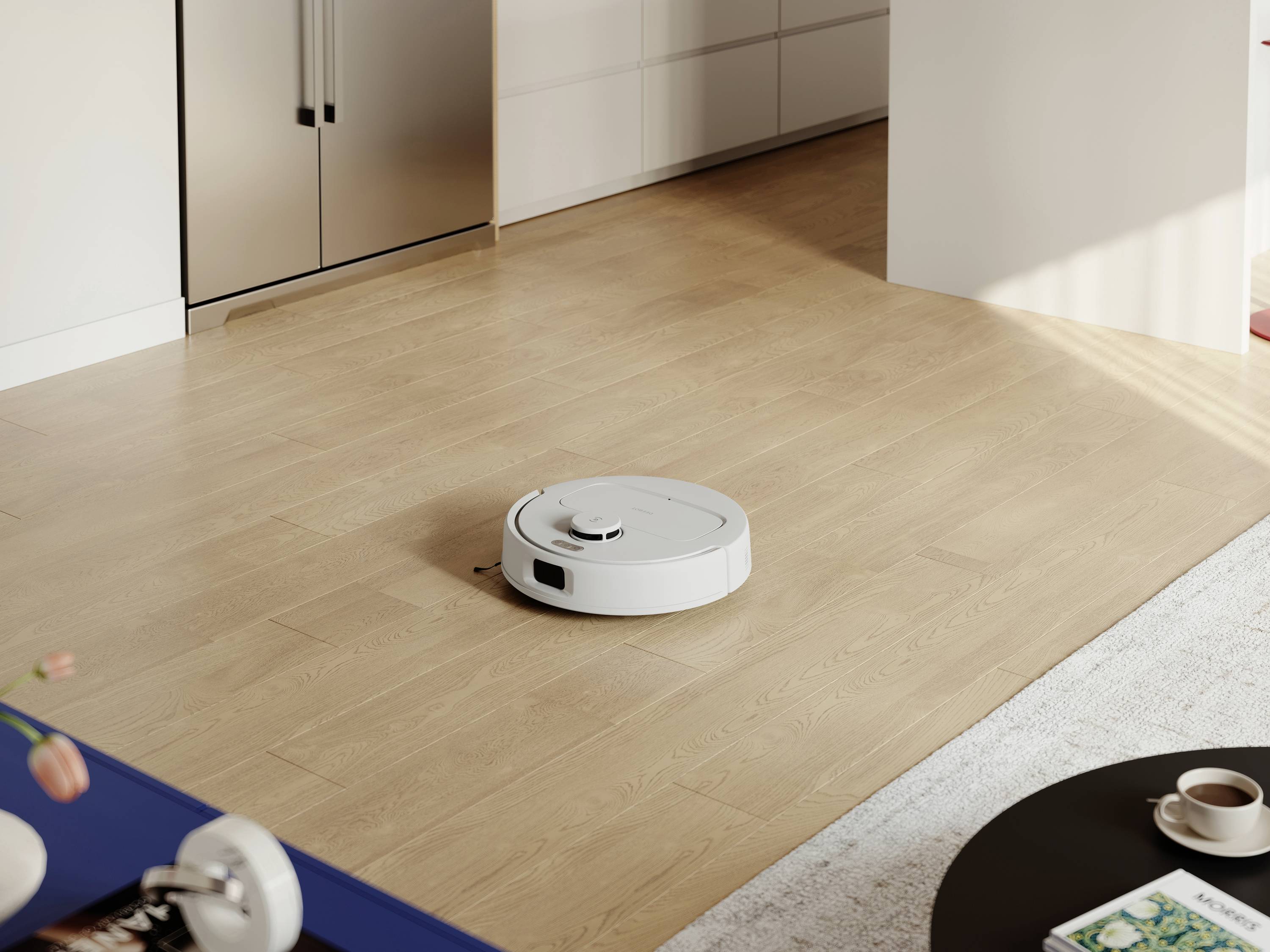 Un robot aspirateur nettoie le sol en bois dans un salon moderne. Des meubles de cuisine sont visibles en arrière-plan.