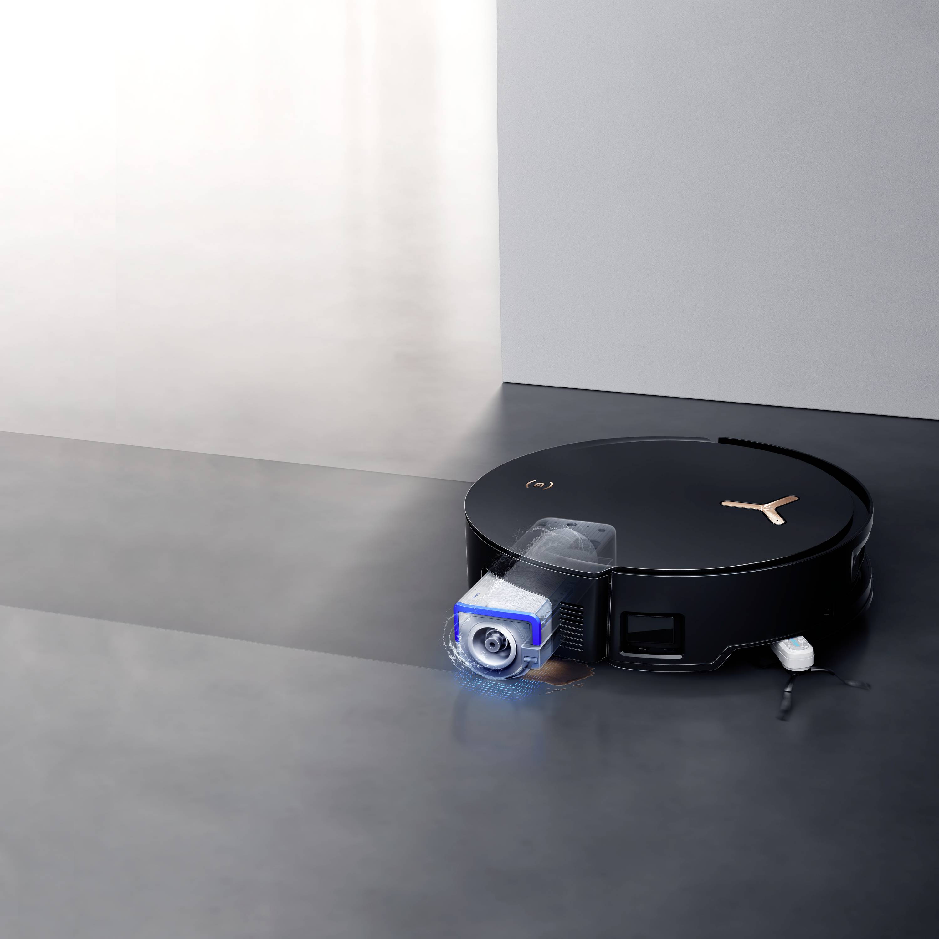 Un aspirateur robot noir avec des touches couleur cuivre nettoie le sol devant un mur gris clair.