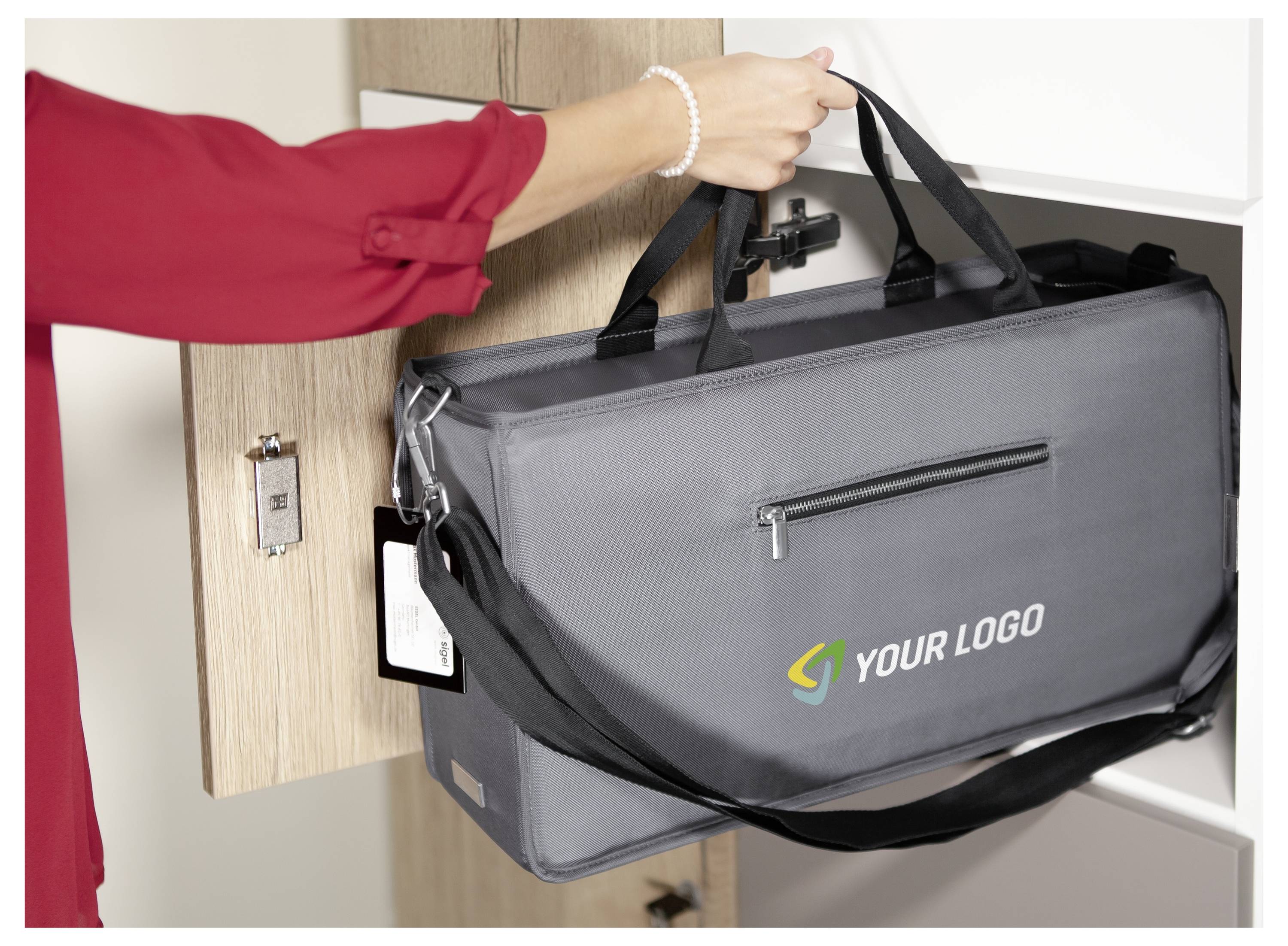 Une personne vêtue d'un chemise rouge retire un sac de livraison isolant gris portant l'inscription « VOTRE LOGO » d'un compartiment fixé au mur.