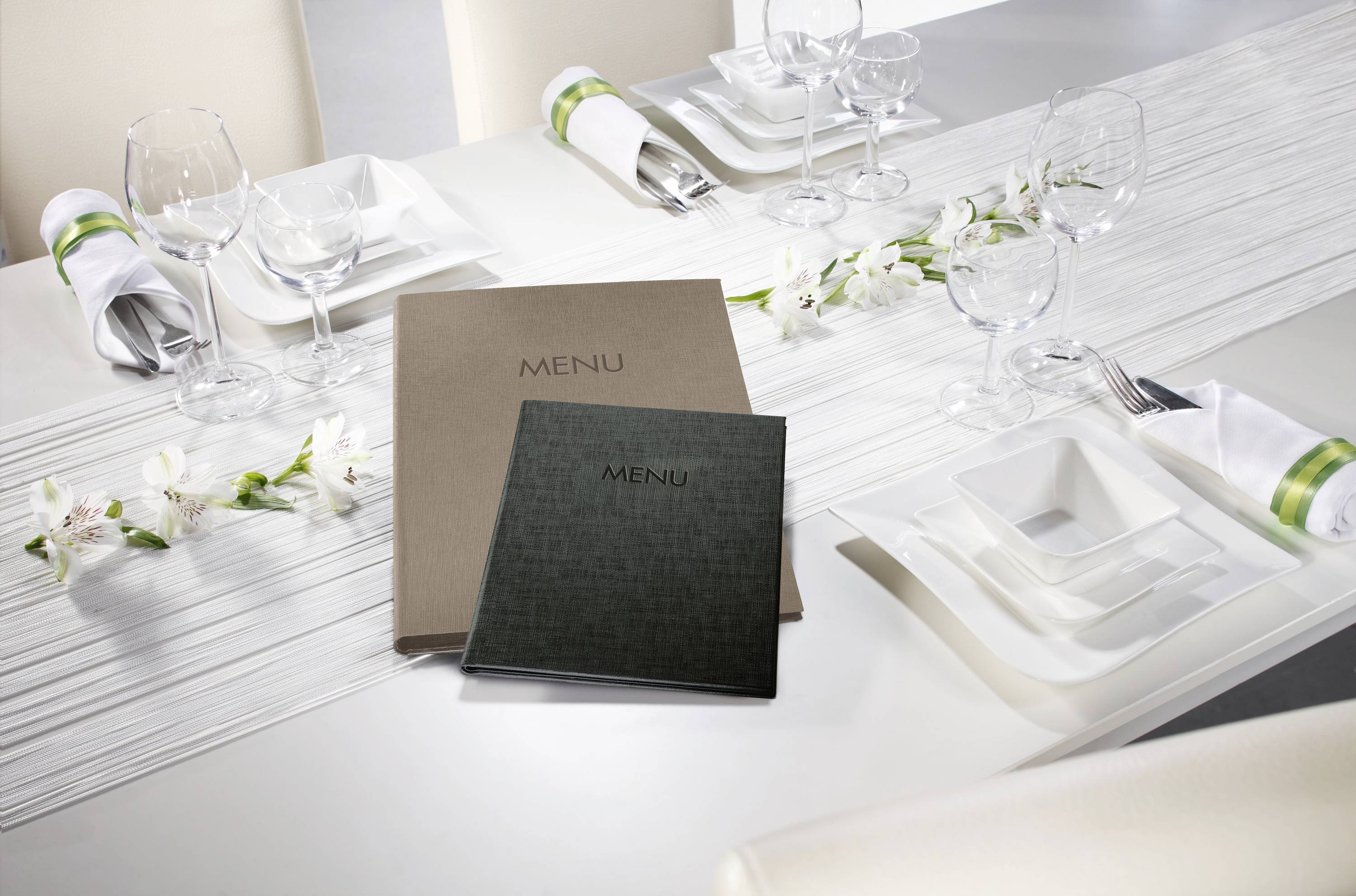 Deux menus sont posés sur une table dressée de blanc, avec des verres à vin, des couverts et des fleurs.