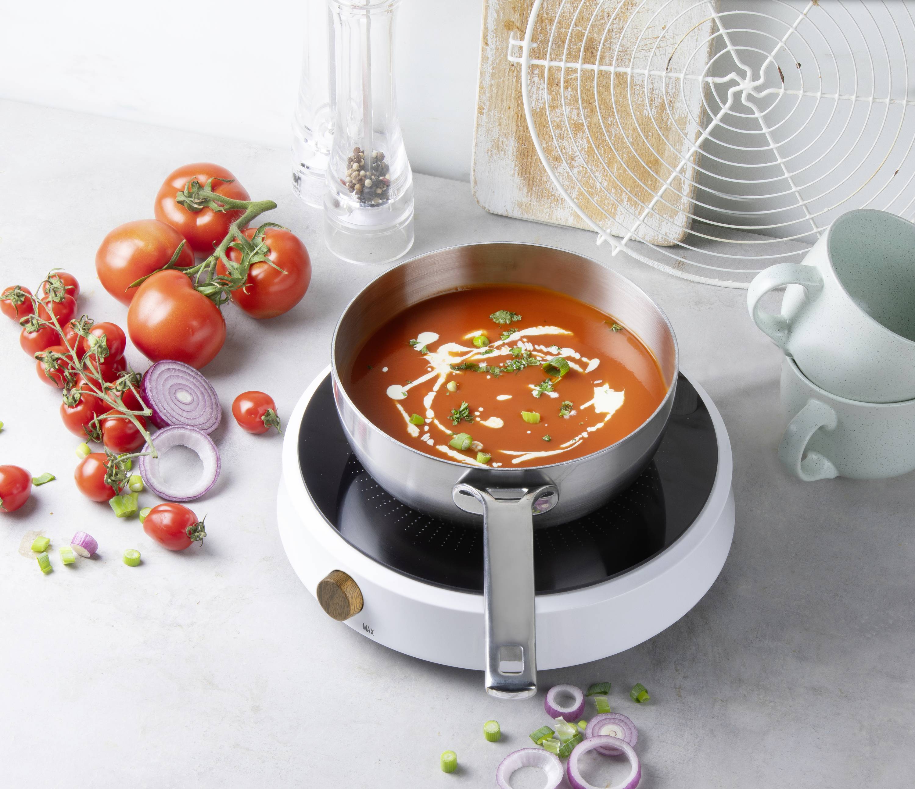 Une casserole de soupe aux tomates sur une plaque de cuisson, entourée de tomates fraîches, d'oignons et d'épices sur une table grise.