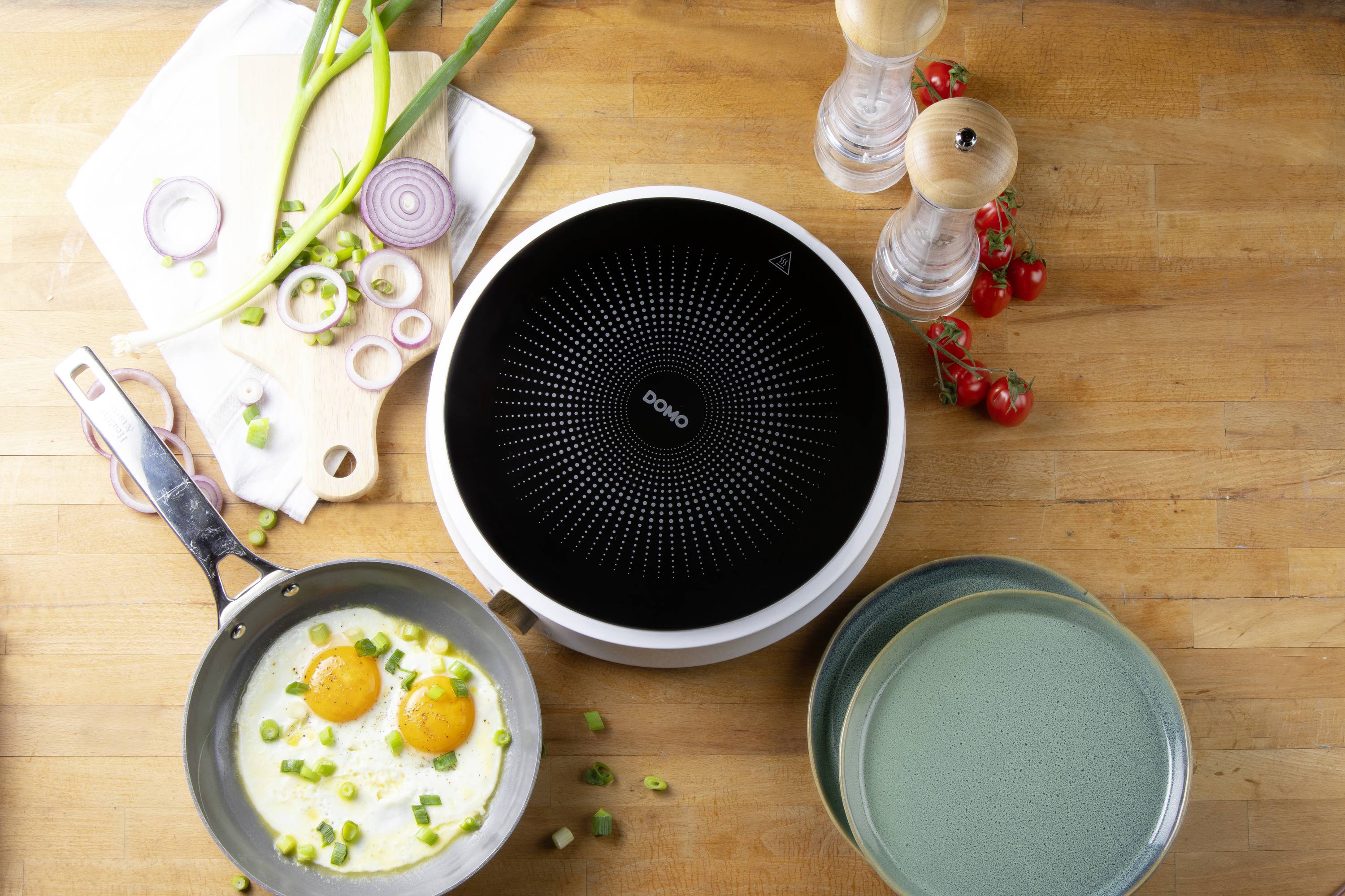 Une poêle avec un œuf au plat, à côté d'oignons, des salières et poivrières, des tomates ainsi que des assiettes sur une planche en bois.