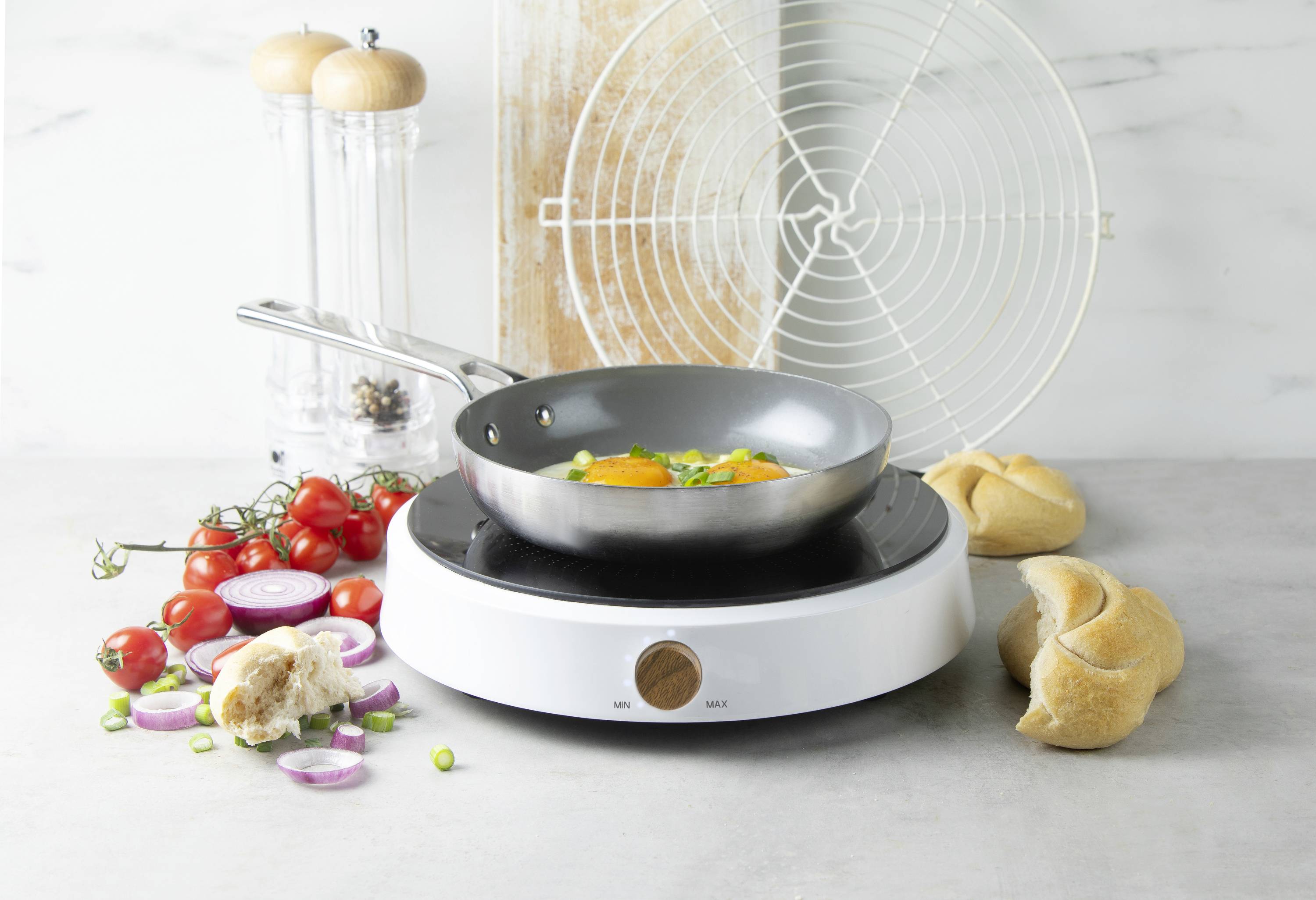 Poêle sur une plaque à induction, avec des œufs au plat et des légumes à l'intérieur. À côté, il y a des tomates, des petits pains, des oignons et un salière et poivrière.