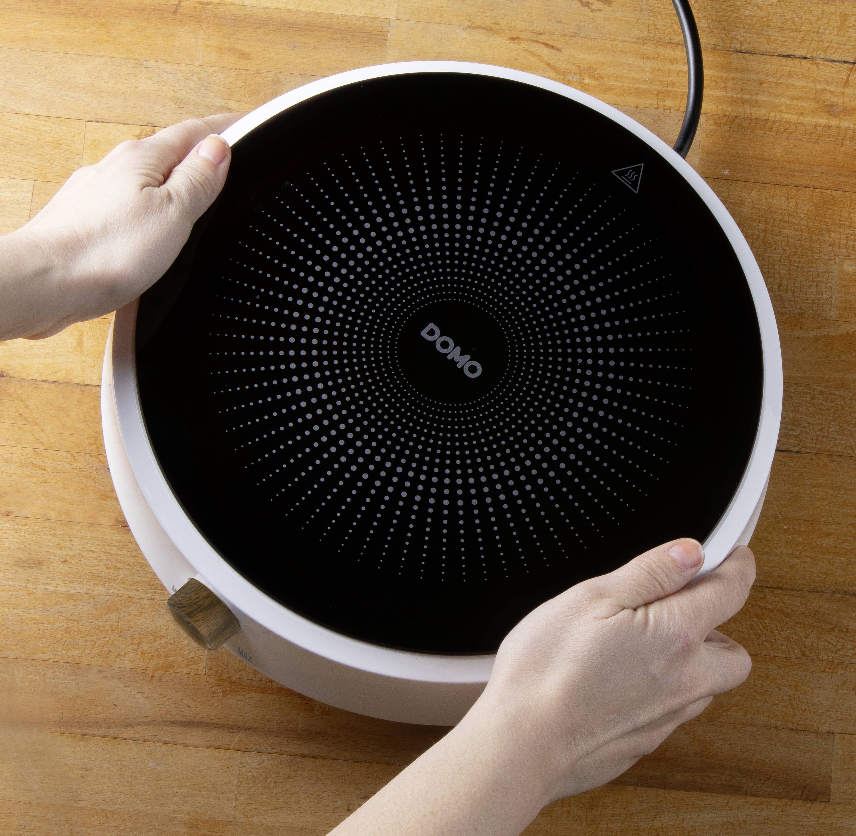 Deux mains tiennent une plaque de cuisson électrique ronde et noire portant l'inscription 'Domo' sur une table en bois.
