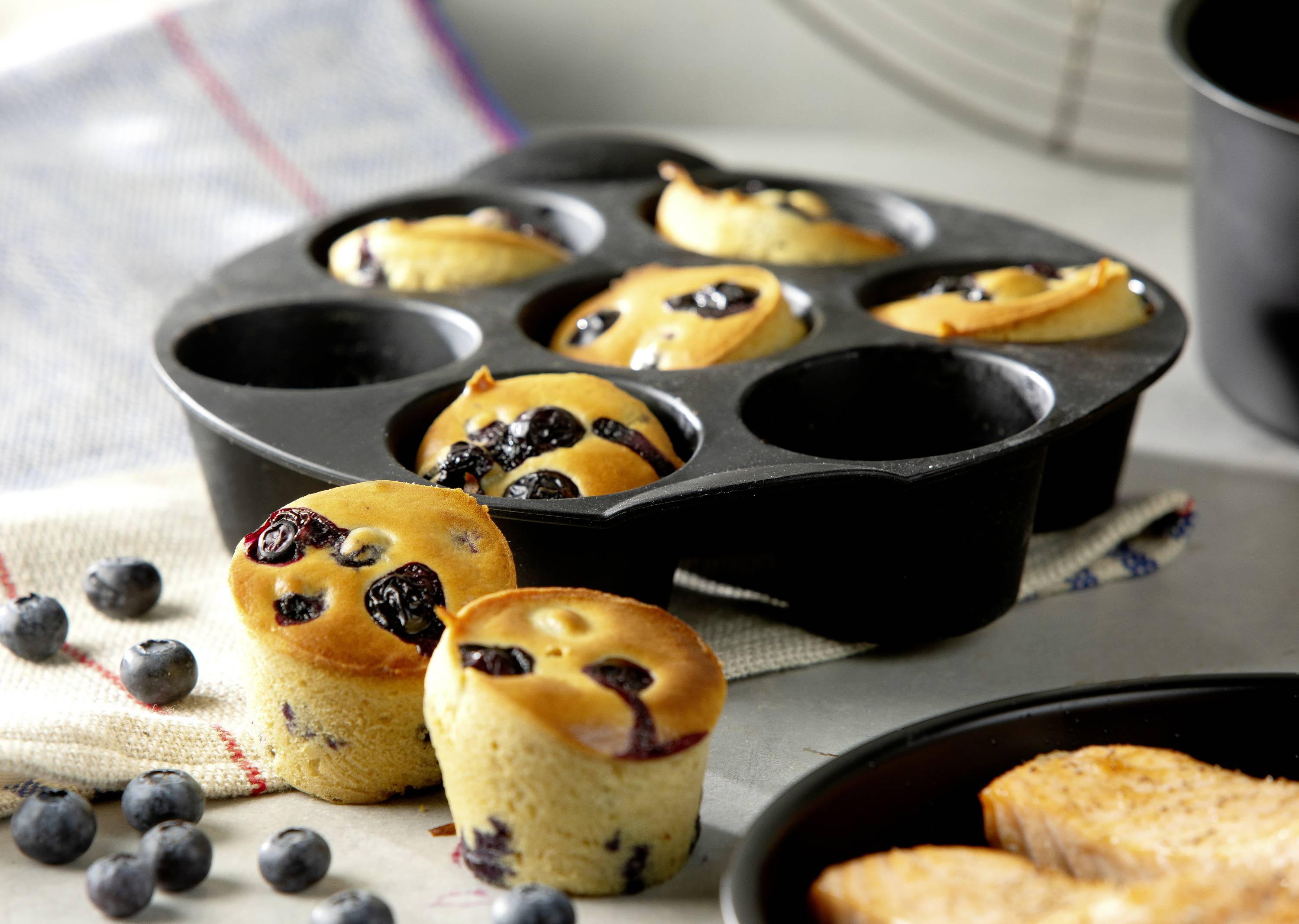 Muffins aux myrtilles dans un moule à pâtisserie noir, quelques-uns posés à côté sur un torchon. Des myrtilles fraîches parsemées autour de cette scène appétissante.
