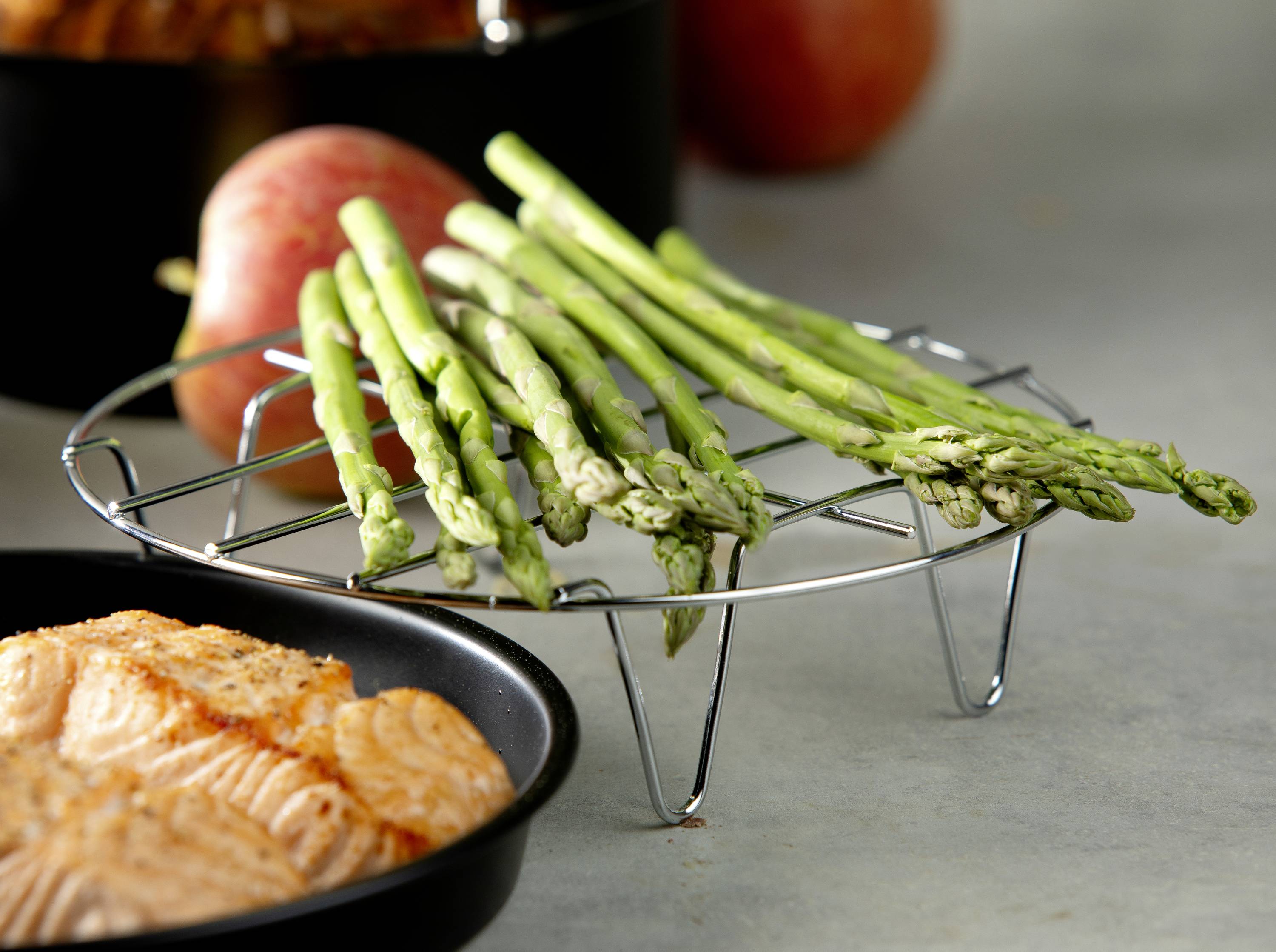 Des asperges sont disposées sur un support métallique rond. Au premier plan, un filet de saumon garni, à l'arrière-plan, une demi-pomme floue.