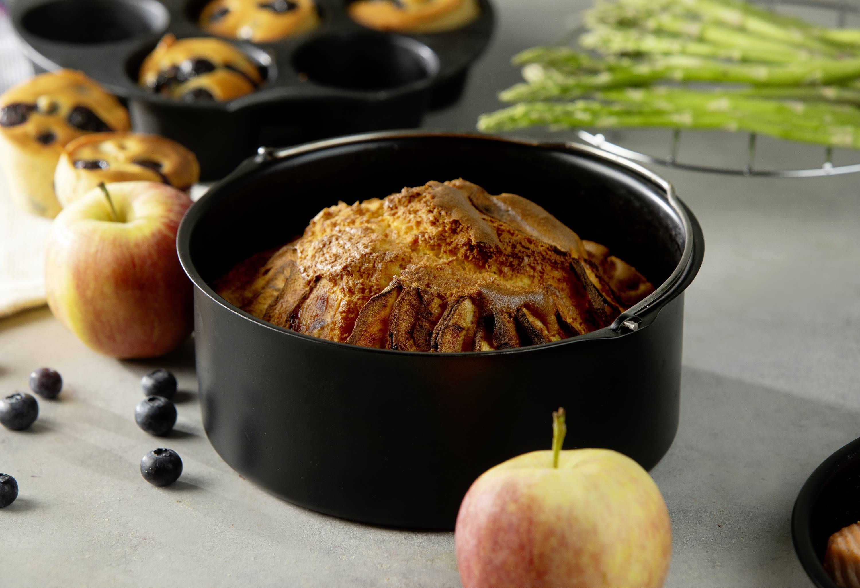 L'image montre un gâteau fraîchement sorti du four dans un moule, entouré de pommes, de myrtilles, de muffins et d'asperges sur une table.