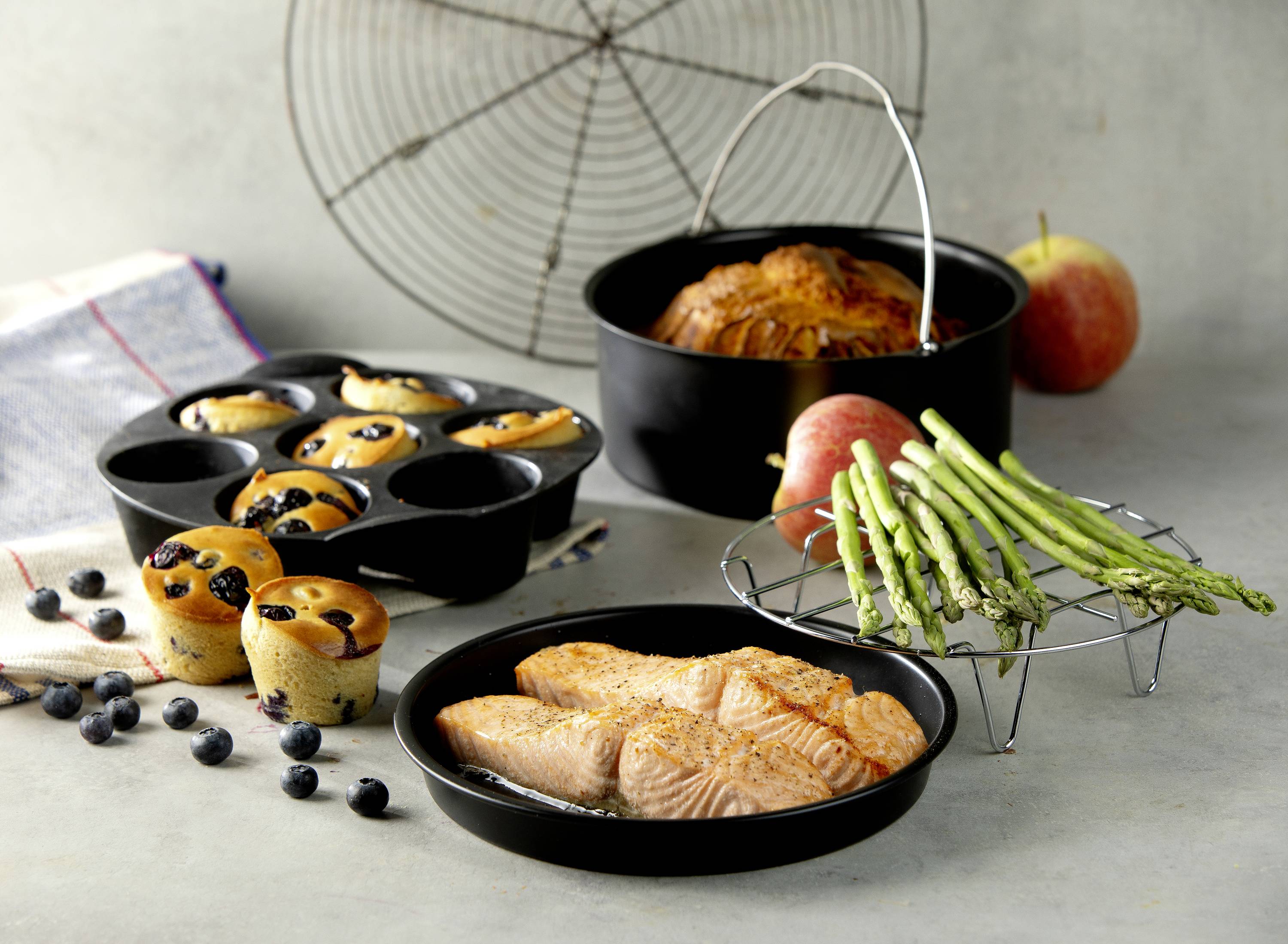 Filets de saumon et asperges sur une assiette, à côté de muffins aux myrtilles et un gâteau rond dans un moule à pâtisserie noir.