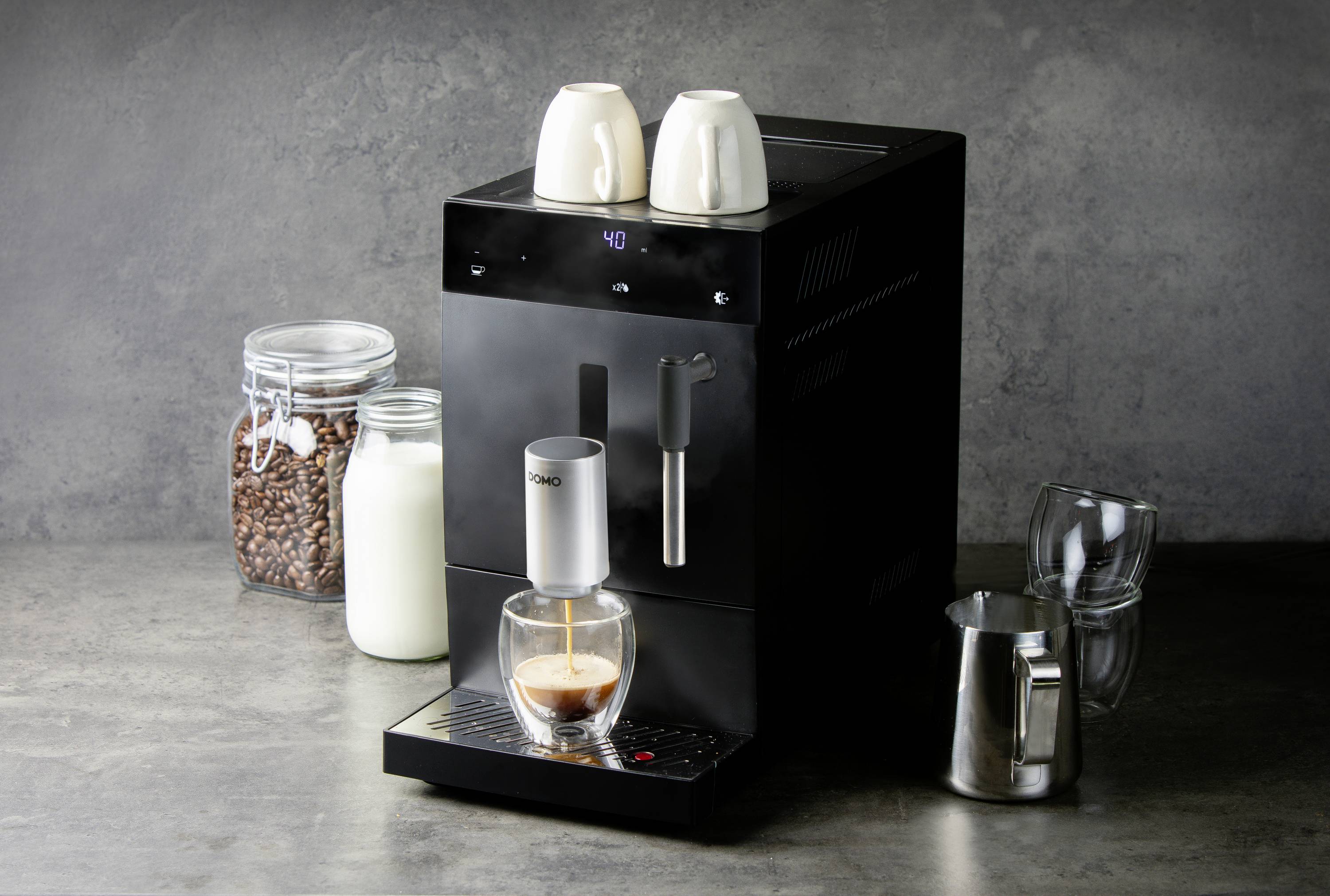 Machine à café noire sur un fond gris, avec des tasses à café en haut, du lait, des grains et des verres à côté. Du café est en train d'être infusé dans un verre.