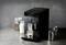 Une machine à café noire prépare un espresso dans un verre. À côté se trouvent un récipient de grains de café et un verre de lait.