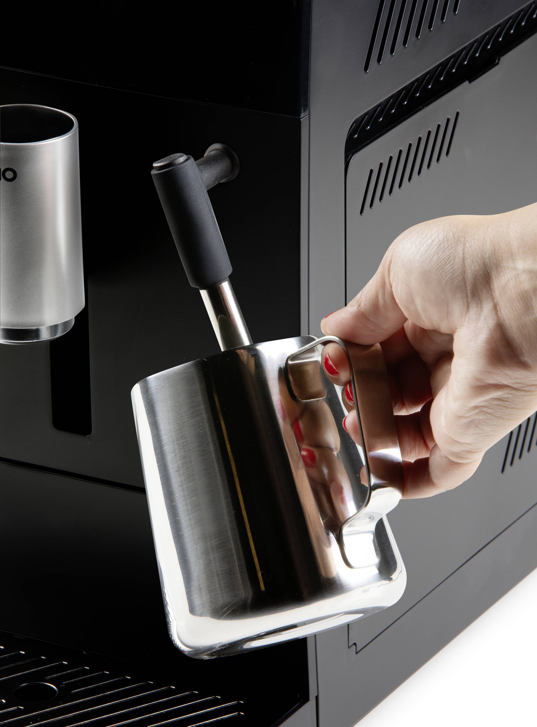 Une main tient un pichet en acier inoxydable sous la buse à vapeur d'une machine à café, prête à faire mousser le lait.