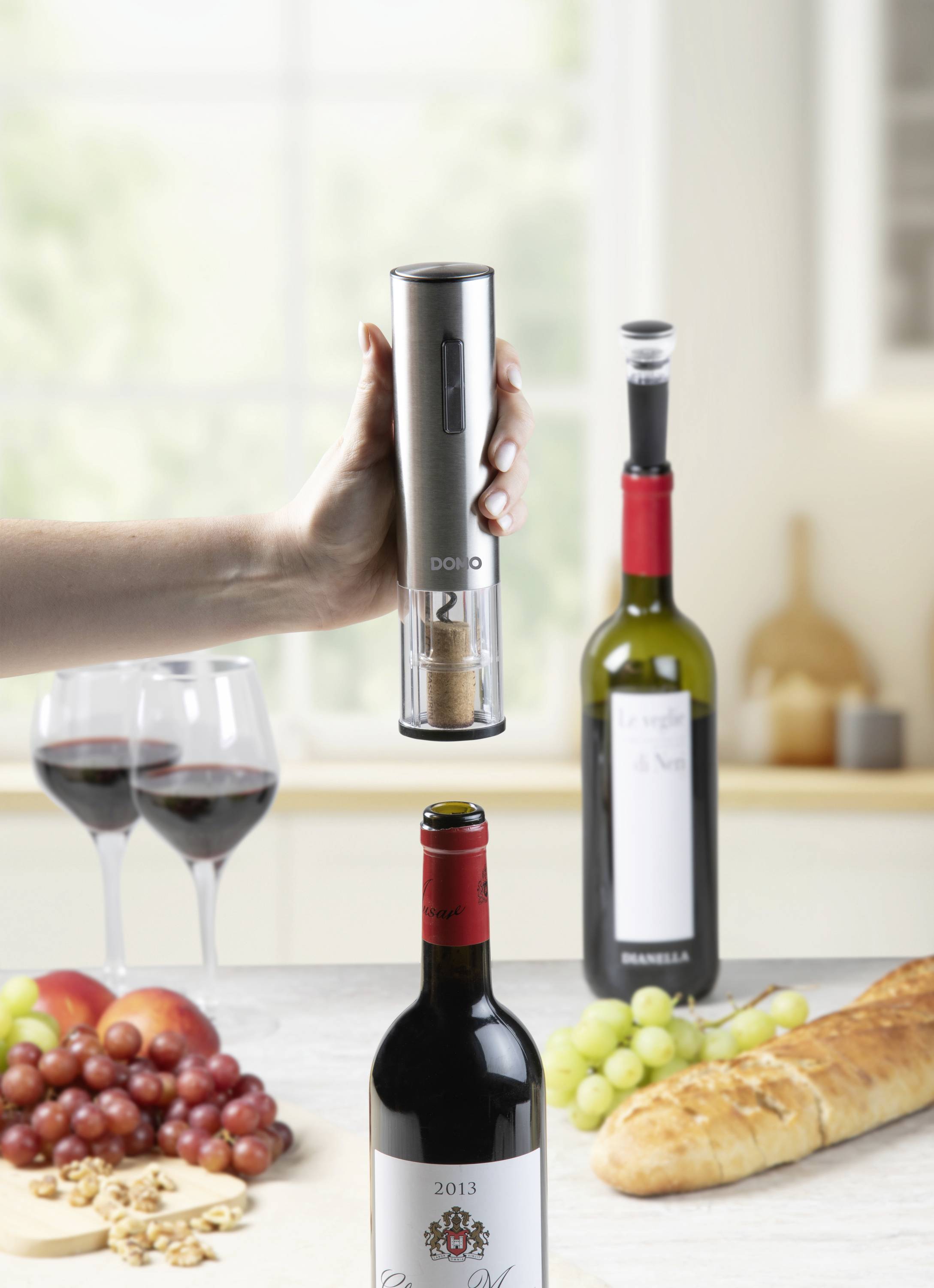 Une main utilise un tire-bouchon électrique pour ouvrir une bouteille de vin. En arrière-plan, deux verres à vin remplis, d'autres bouteilles de vin, des raisins et un baguette sont posés sur une table.