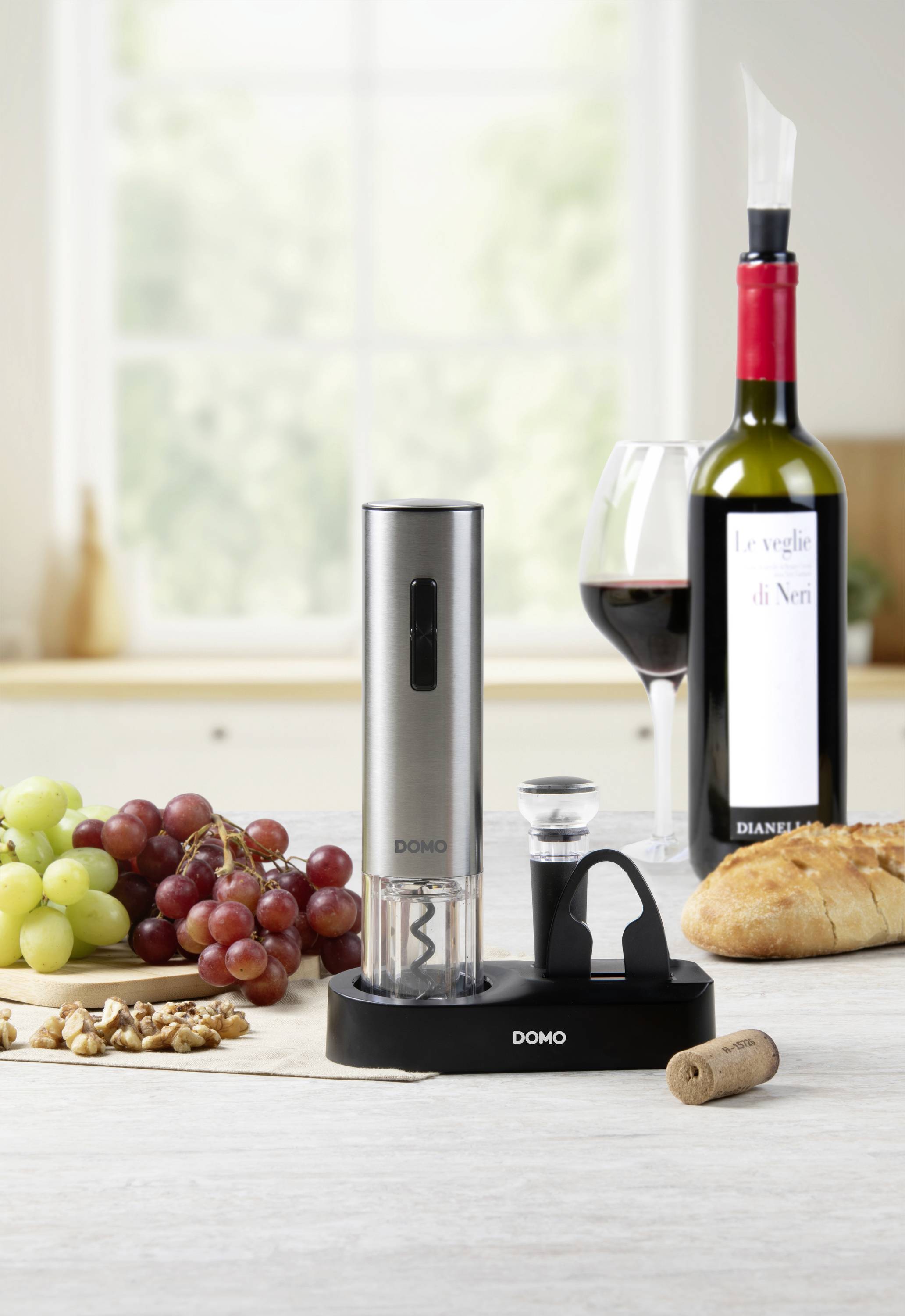 Un tire-bouchon électrique avec une bouteille de vin, un verre de vin rouge, des raisins, des noix et du pain sur une table près d'une fenêtre.