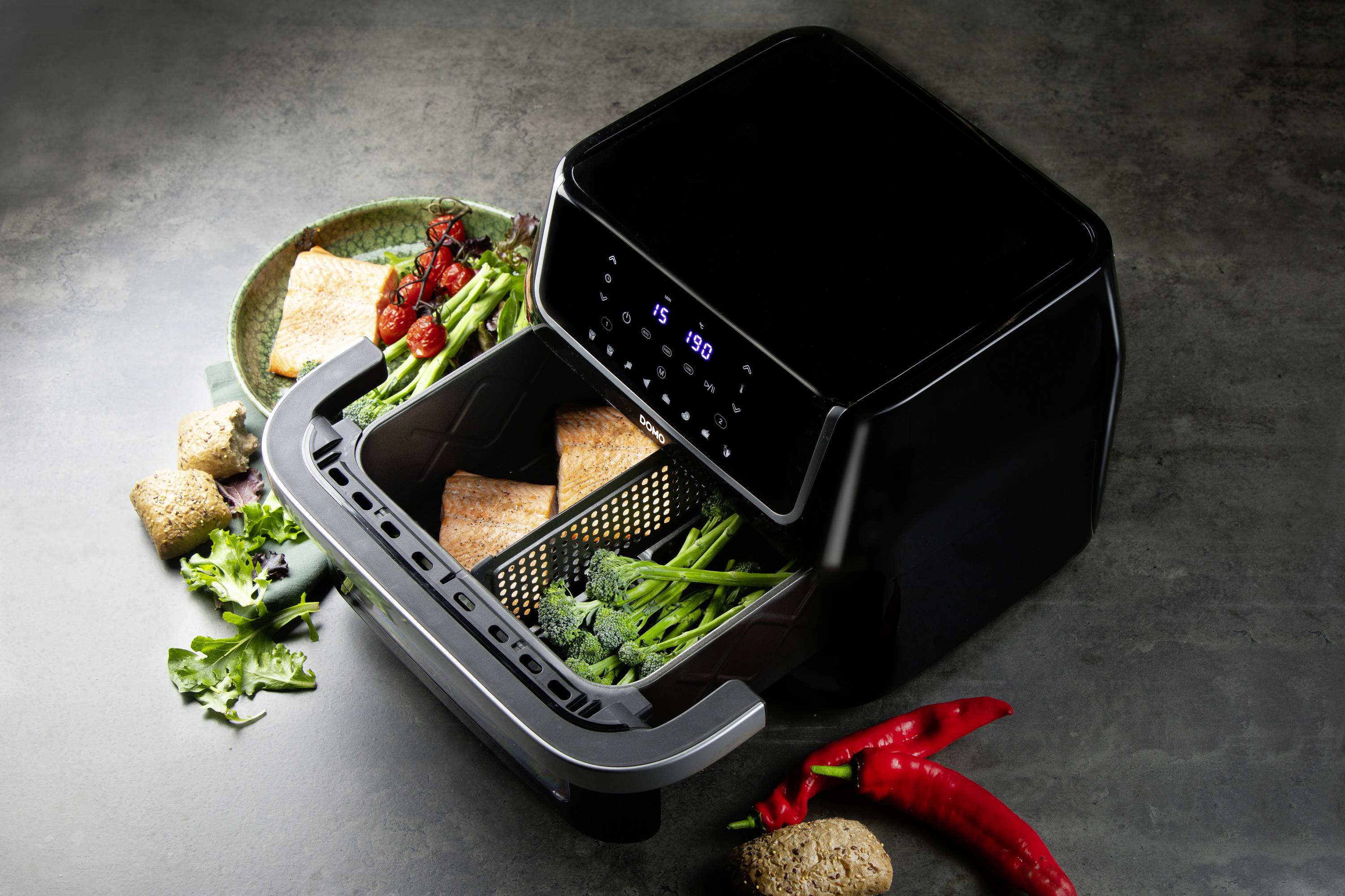 Friteuse à air chaud avec affichage numérique, remplie de brocoli et de tofu. À côté, des légumes frais sur une assiette et des piments.