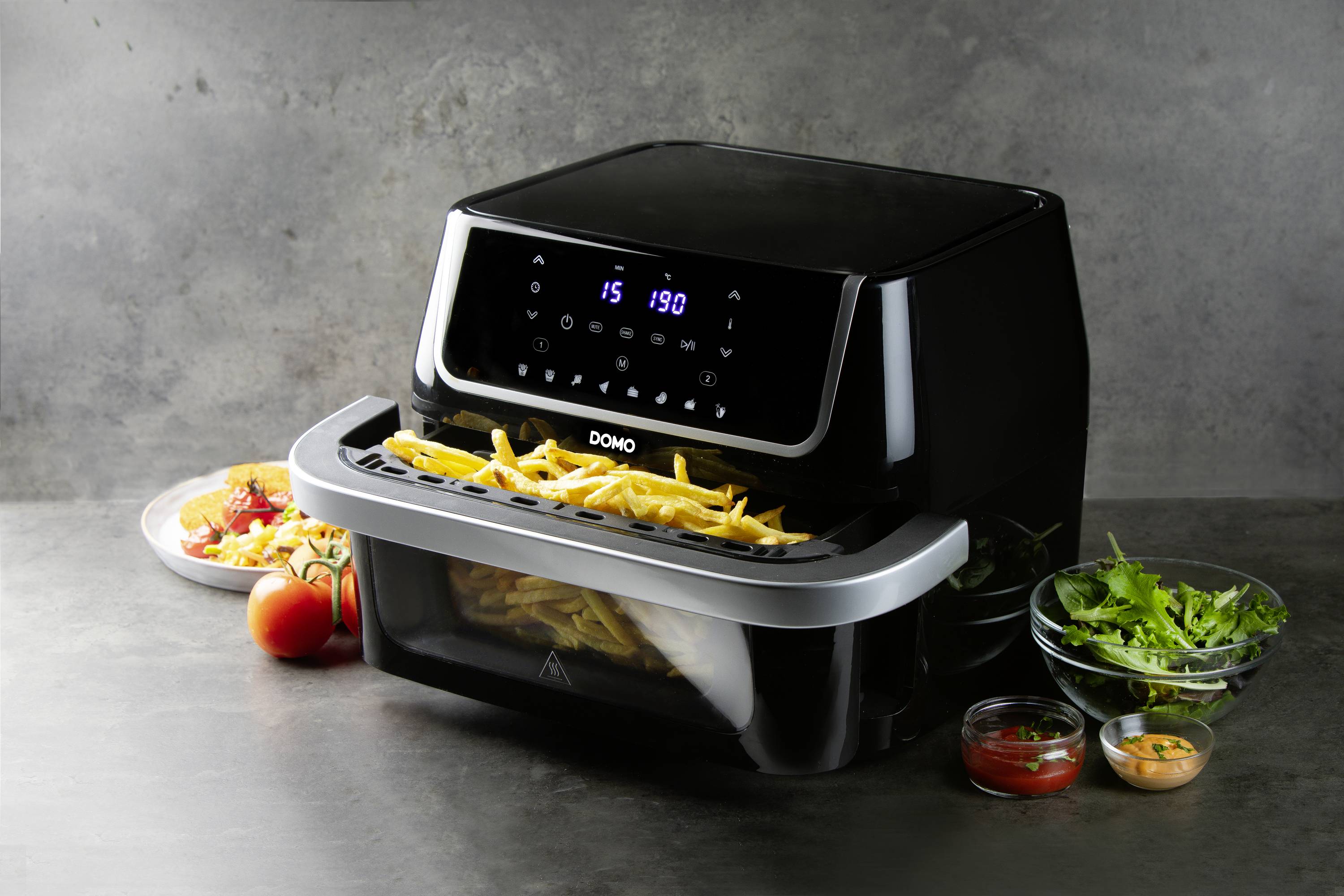 Une friteuse à air chaud sur une table grise, remplie de frites. À côté se trouvent des assiettes avec de la salade, des tomates et des ramequins de sauce.