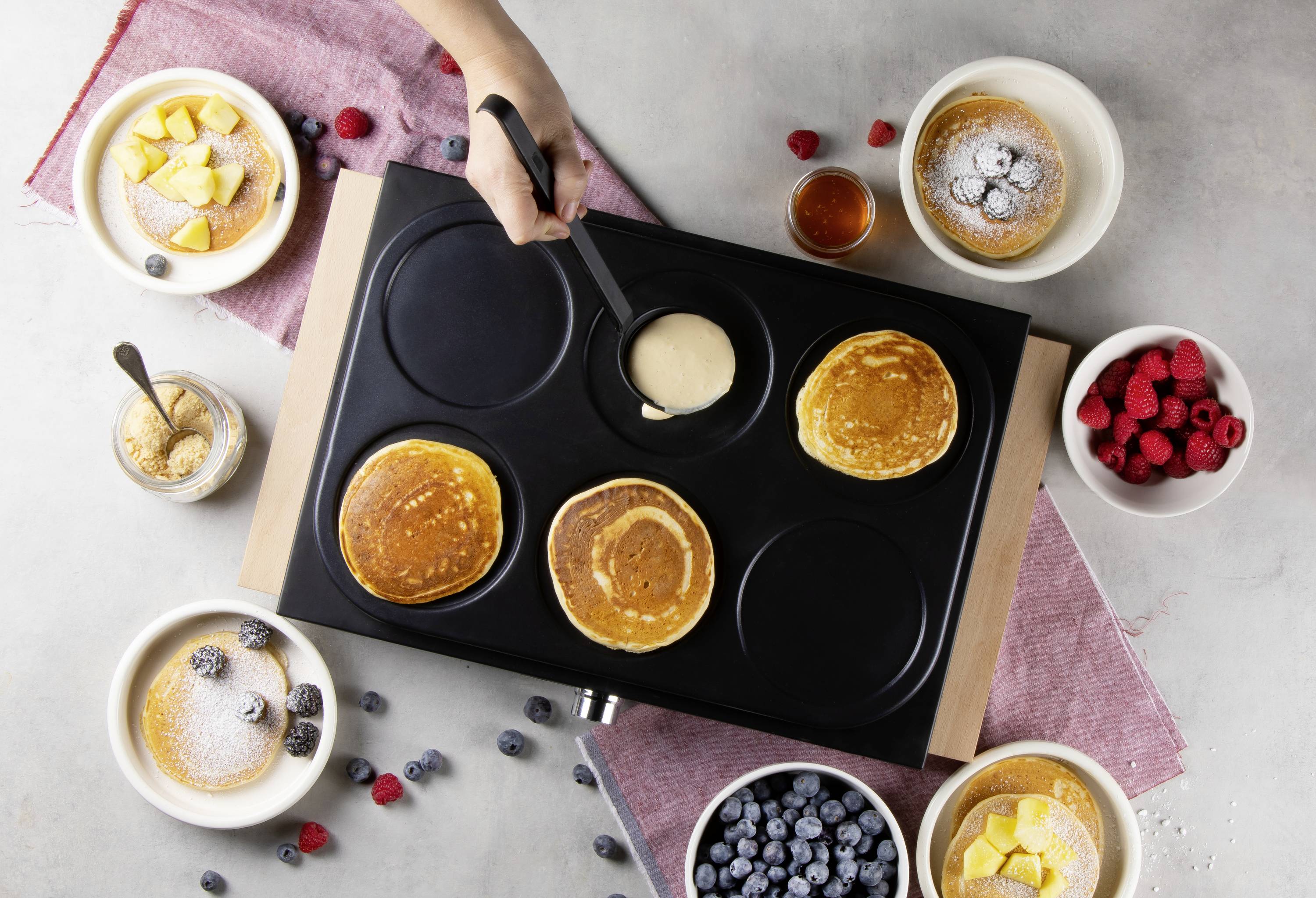 Une table avec une poêle où des pancakes sont en train de cuire. À côté, des bols contenant des myrtilles, des framboises, du sucre glace et du sirop.