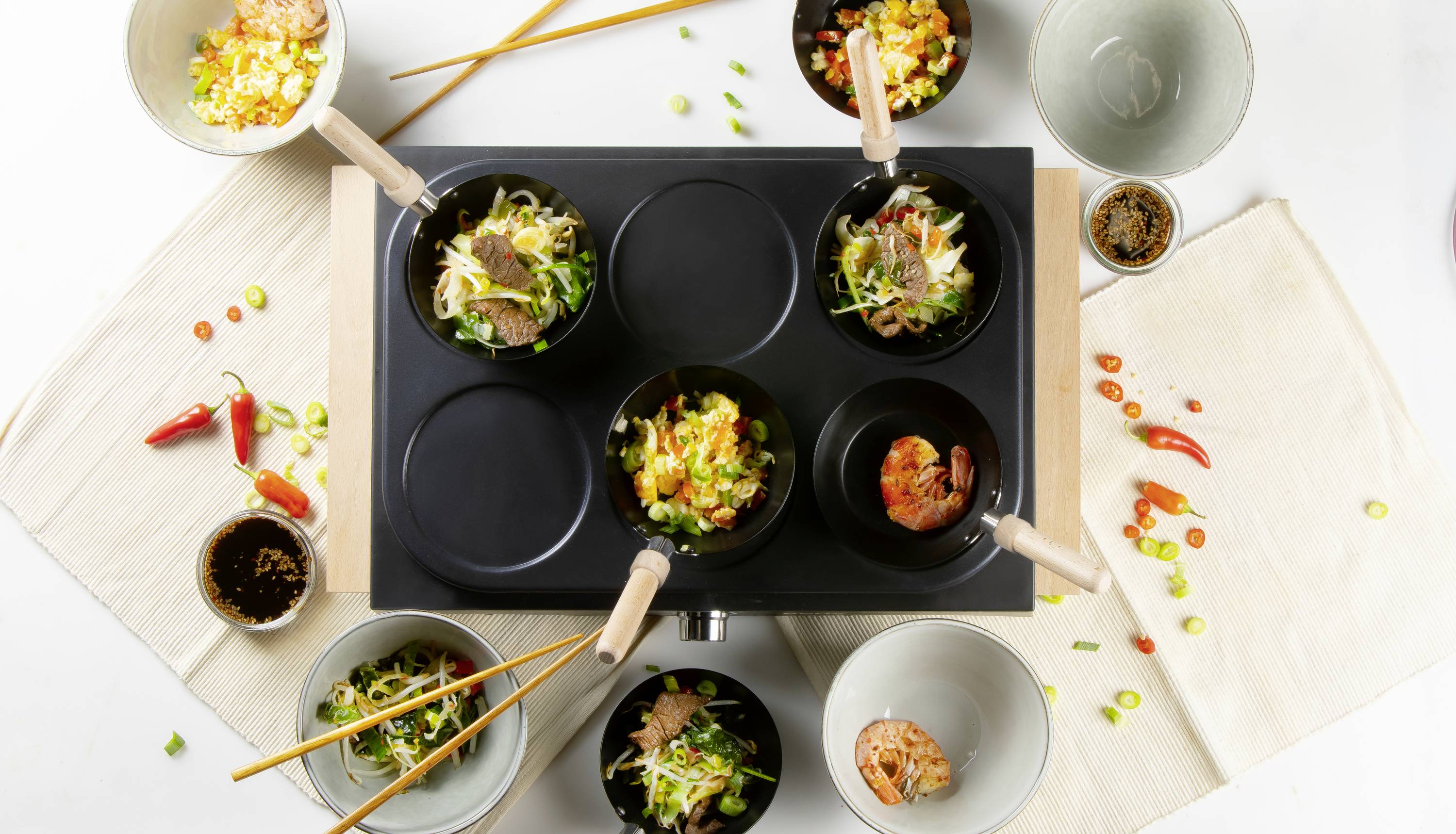 Cinq poêles contenant différents plats asiatiques sont disposées sur une cuisinière, entourées de bols, de baguettes et d'épices.