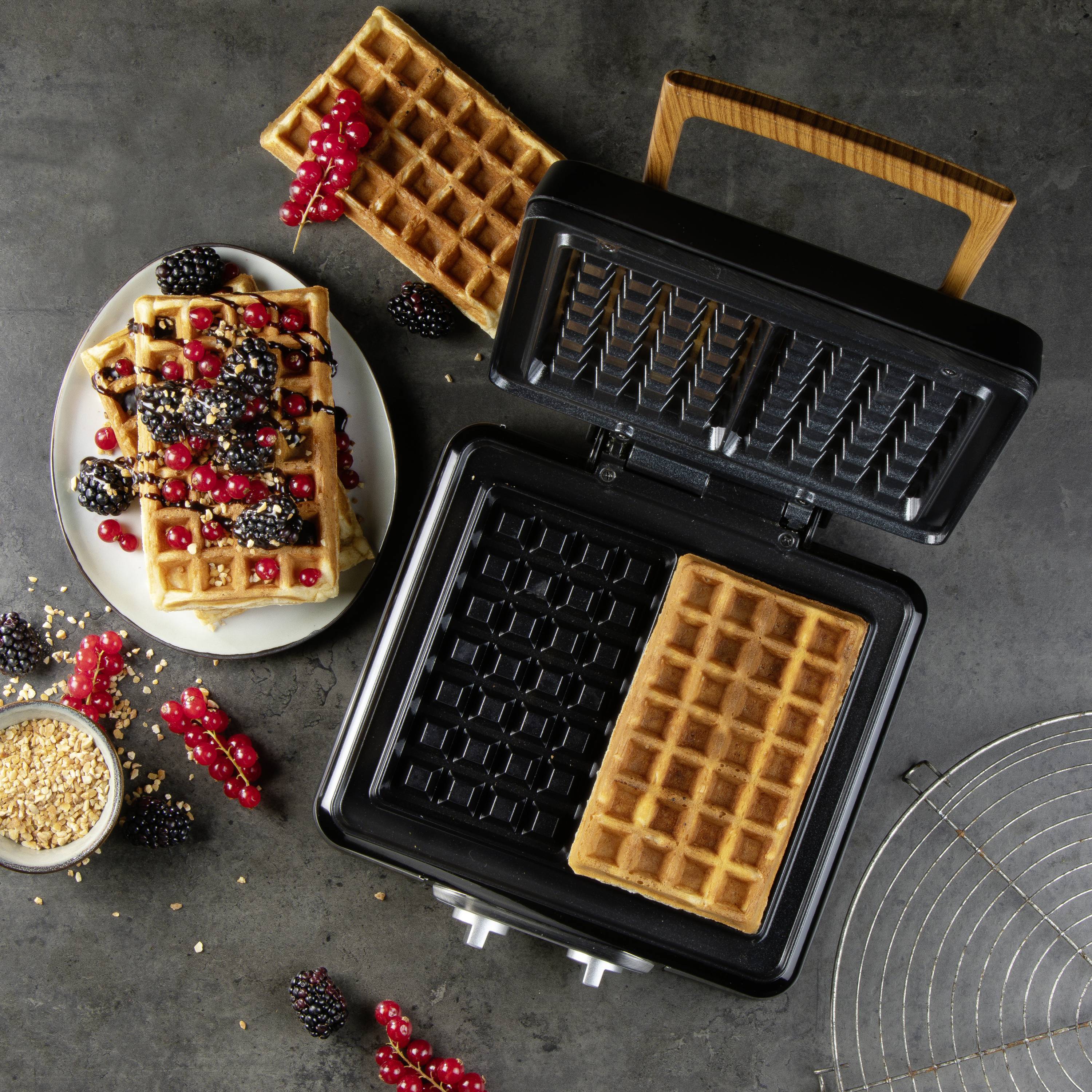 Un gaufrier avec une gaufre, entouré de baies fraîches et d'une assiette de gaufres garnies.