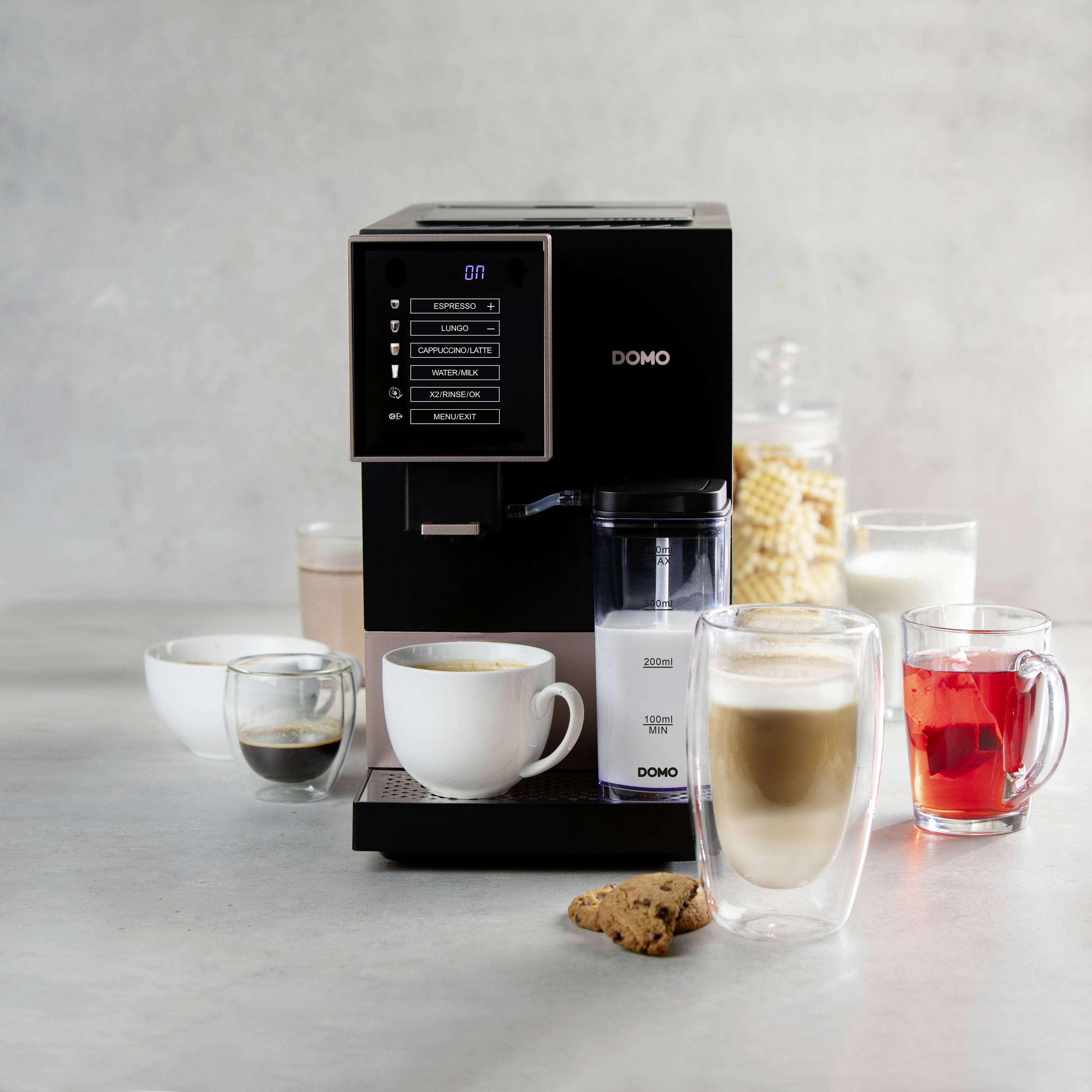 Machine à espresso avec réservoir à lait, trois verres de boissons et un biscuit sur la table. Les boissons sont un espresso, un café au lait et un thé.